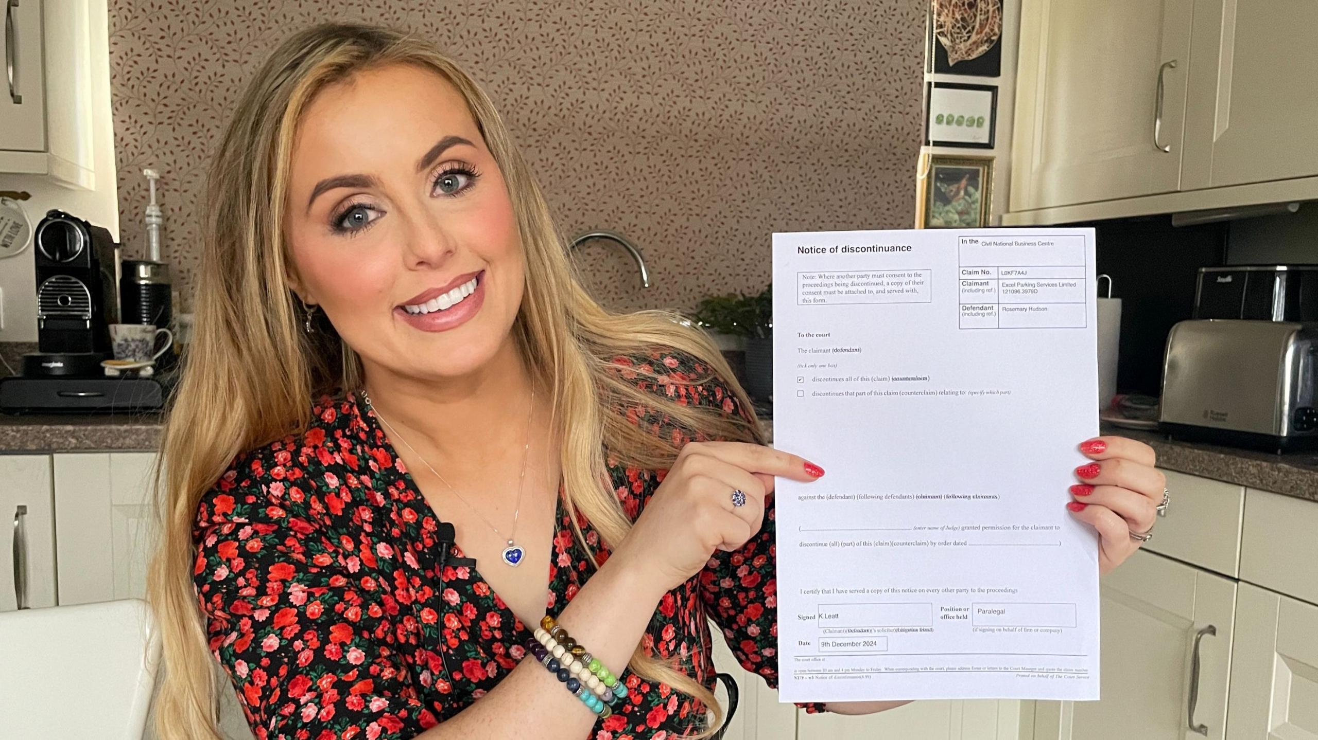 Rosey Hudson holds up her notice of discontinuance while sitting at her kitchen table, she's smiling and wearing a floral red top with red painted fingernails