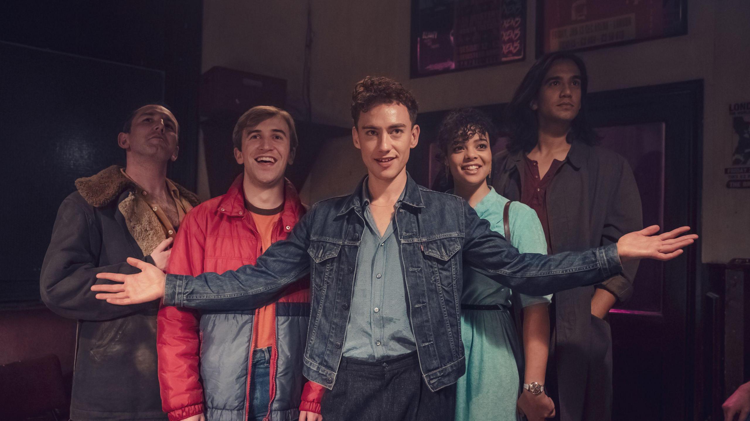 This photograph includes five friends. From left to right we have a man in a black jacket with a fur collar, a man in a red puffer coat, a man in a denim jacket, a woman in a blue dress and finally (on the far right) a man in a grey jacket. They're all looking in different directions.