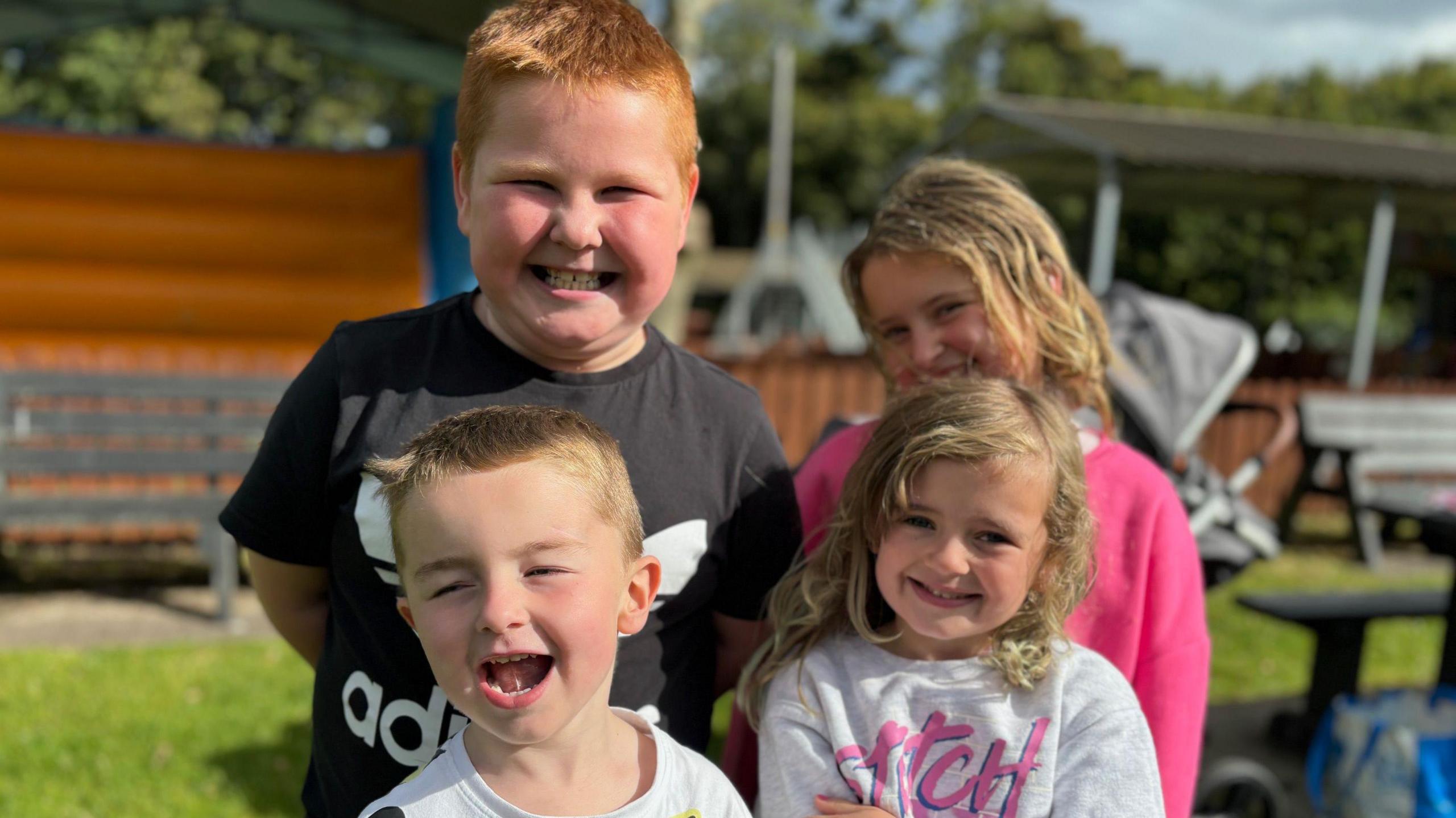 Four children smiling at the camera - there are two smaller ones at the front, a blond boy and girl. There is a boy with red hair grinning at the camera in the back row, with another curly-haired blond girl beside him.