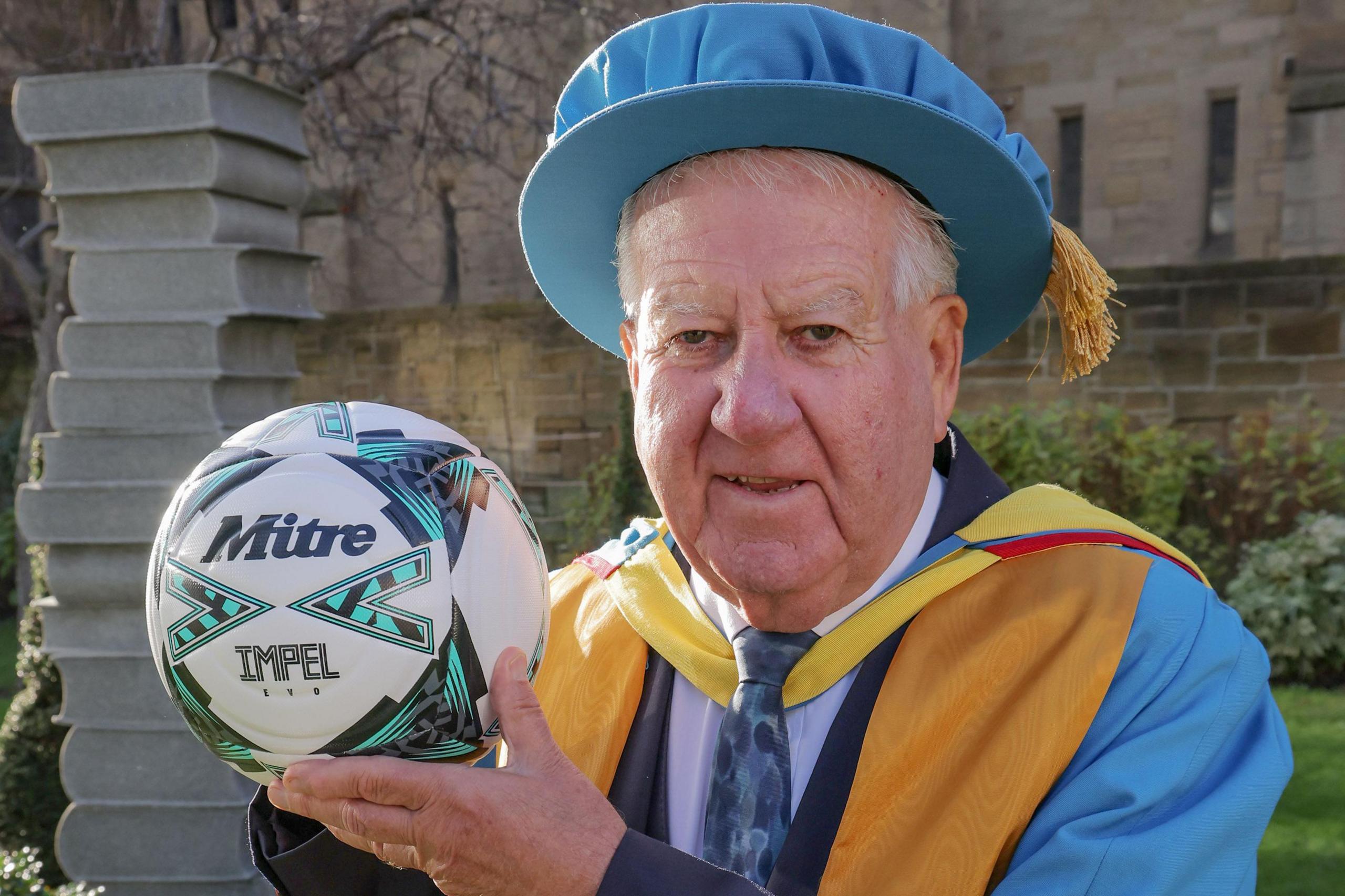 Sir Bob Murray wearing a bright blue cap and gown. He is wearing a blue tie and holding a white and blue "Mitre" ball. He is smiling and posing at the camera. There is greenery in the back and a statue of a stack of books. 