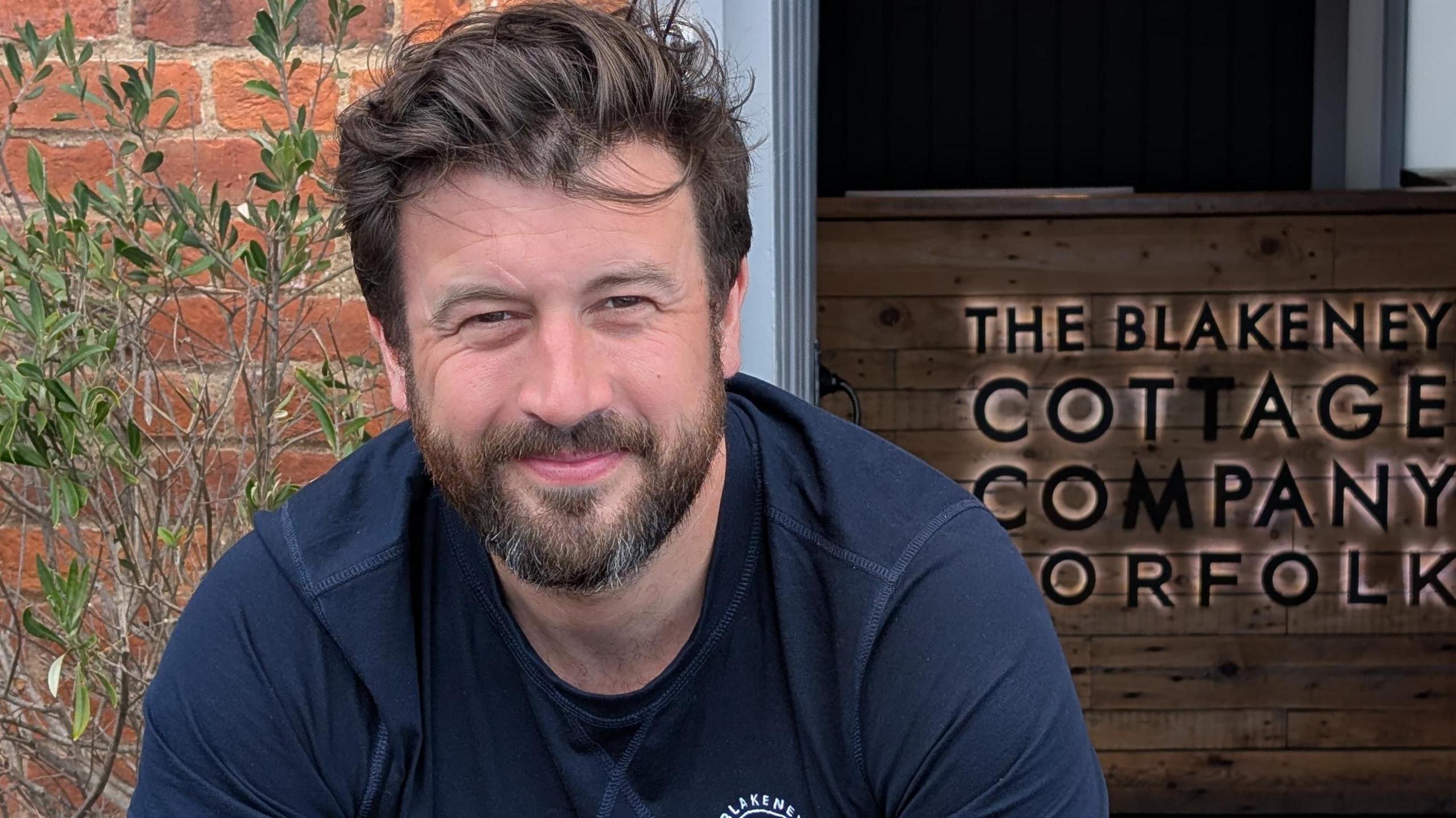 Man smiling into the camera wearing a dark blue jumper in front of a sign saying the Blakeney Cottage Company