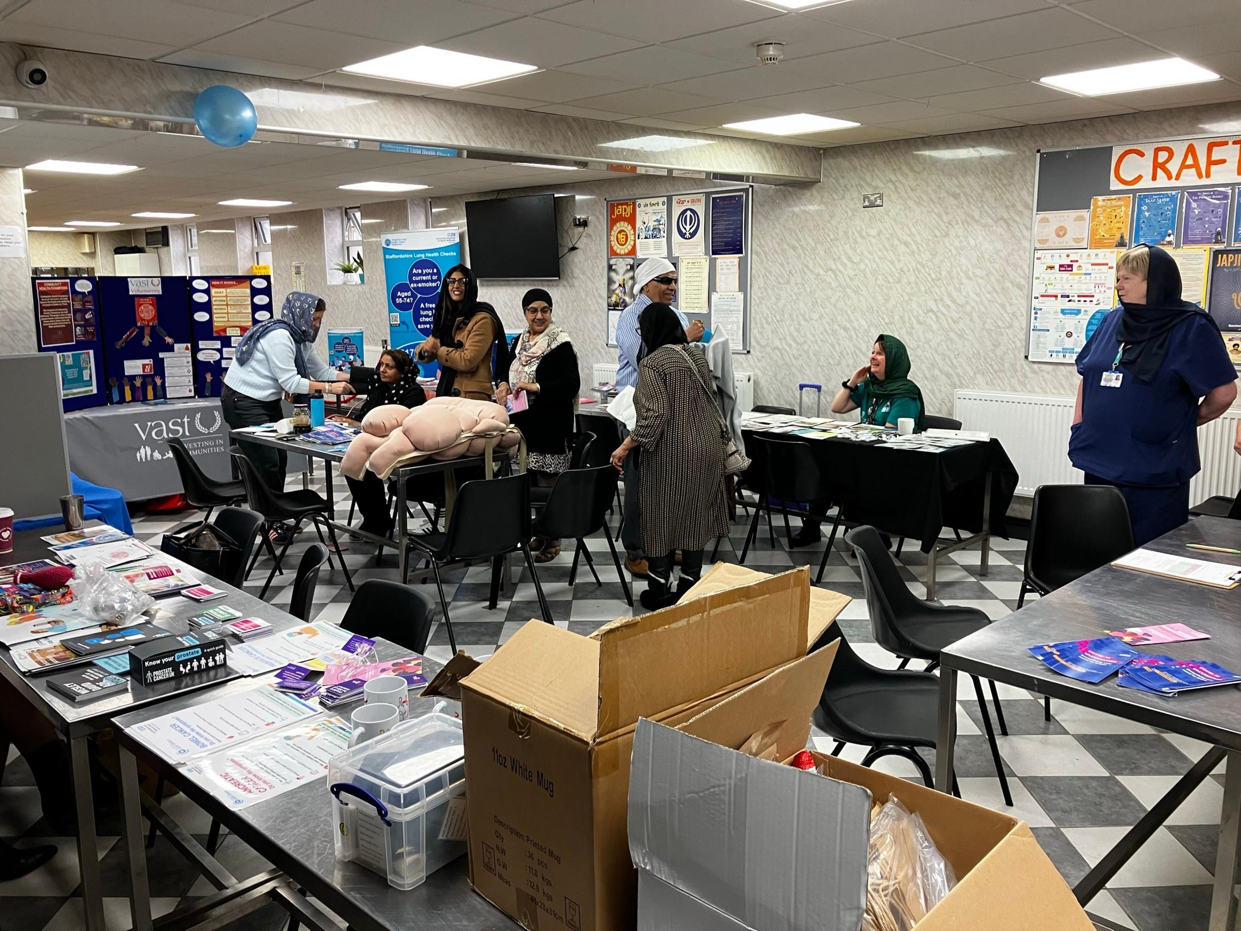 Several people in a room in the gurdwara with lots of tables and chairs.