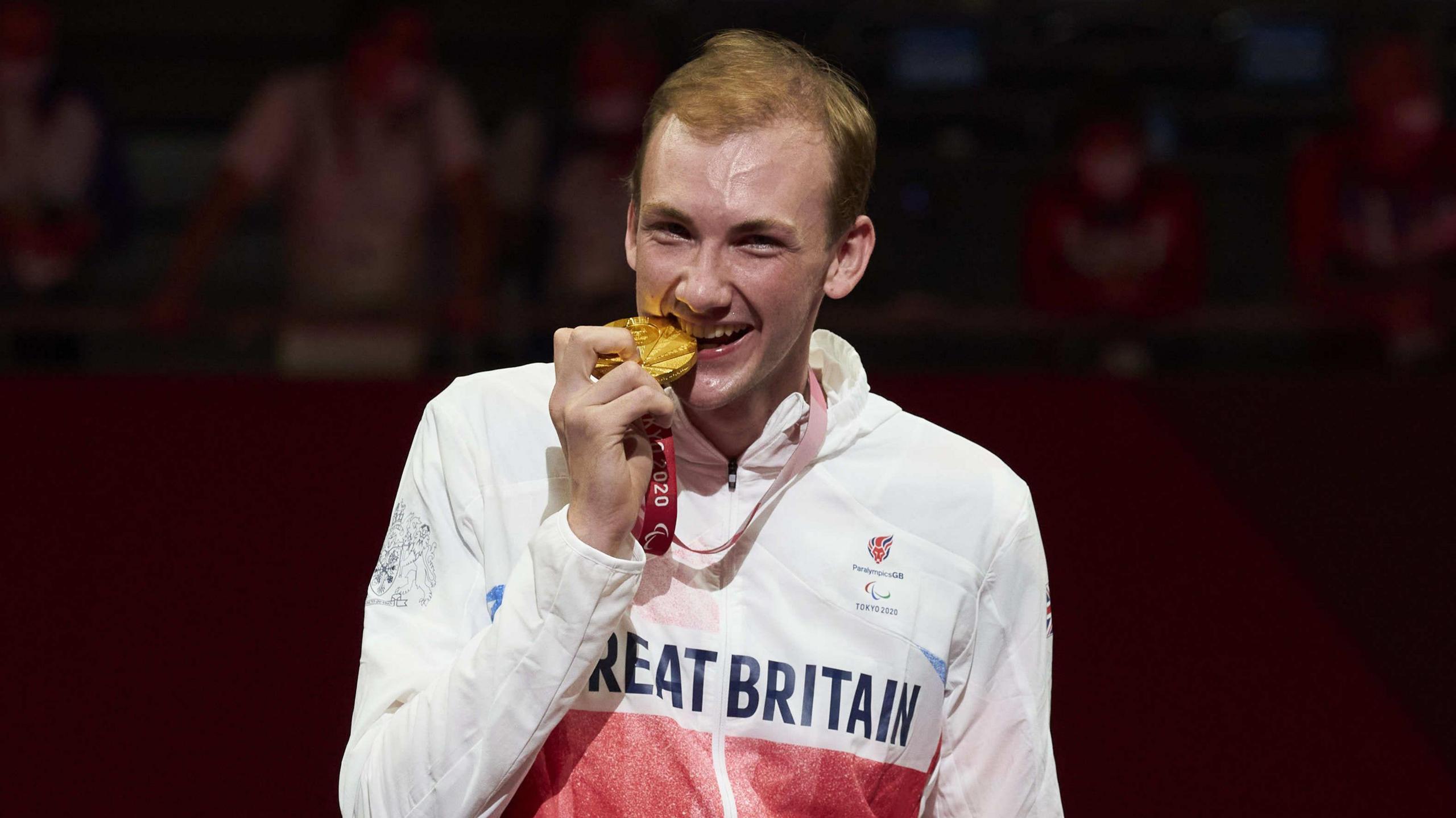 Fencer Piers Gilliver smiles as he pretends to bite a gold medal he has won at the Paralympics in Tokyo. He is wearing a white top with the words Great Britain on it