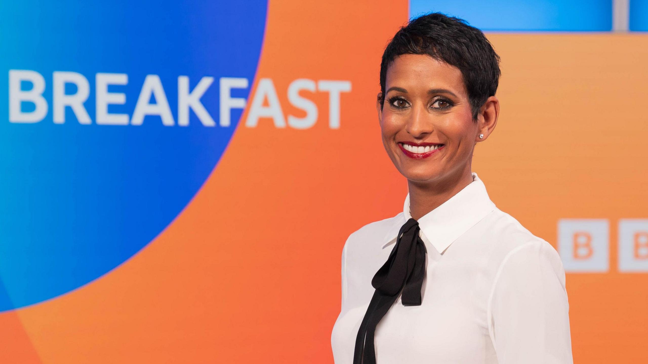 Naga Munchetty posing and smiling in front of the BBC Breakfast studio and logo