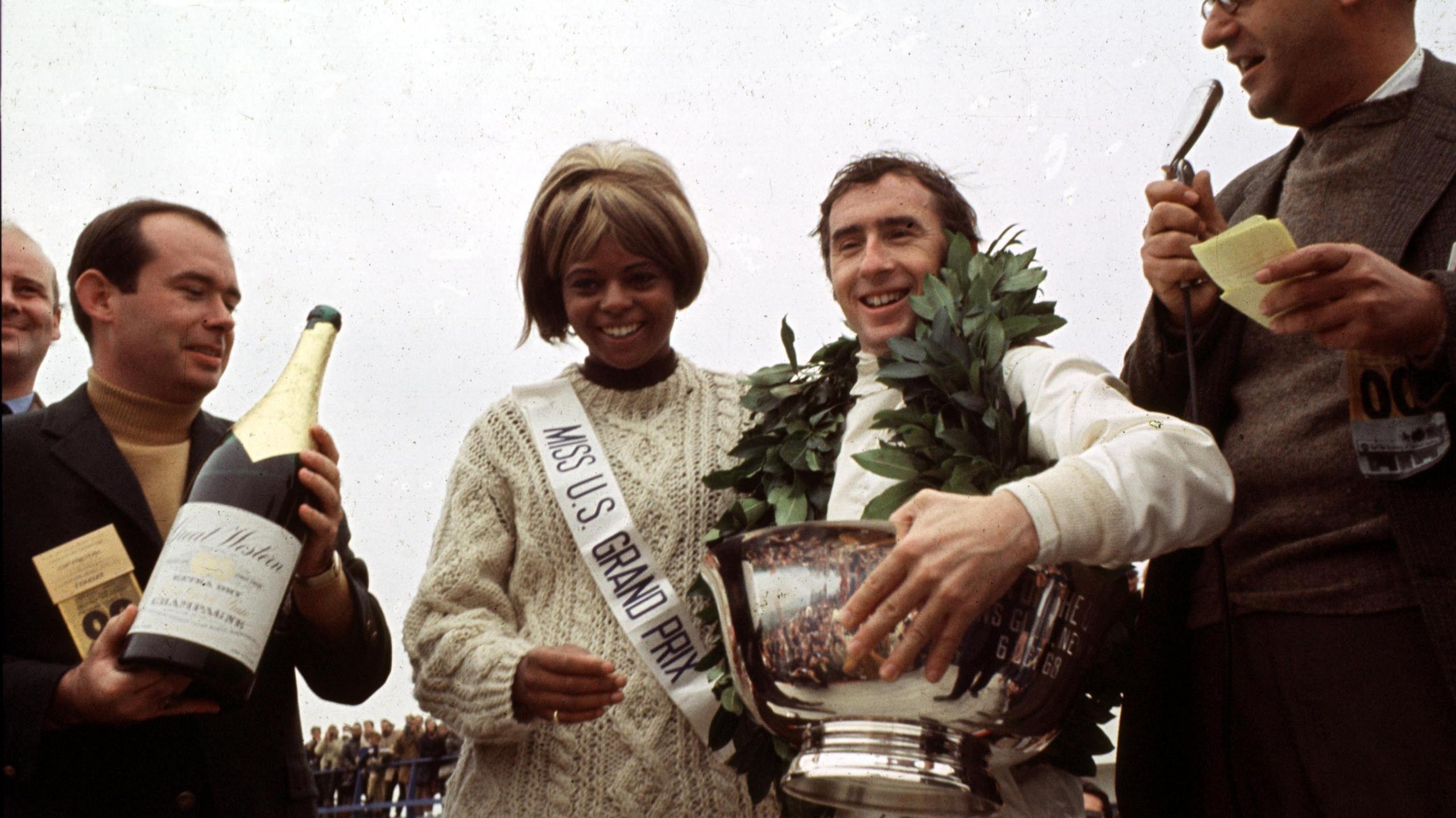 Race winner Jackie Stewart holds the winner's trophy while wearing a laurel around his neck after the 1968 United States Grand Prix. Stood around him are an interview, a man holding a bottle of champagne and a woman wearing a sash that says 'Miss US Grand Prix'