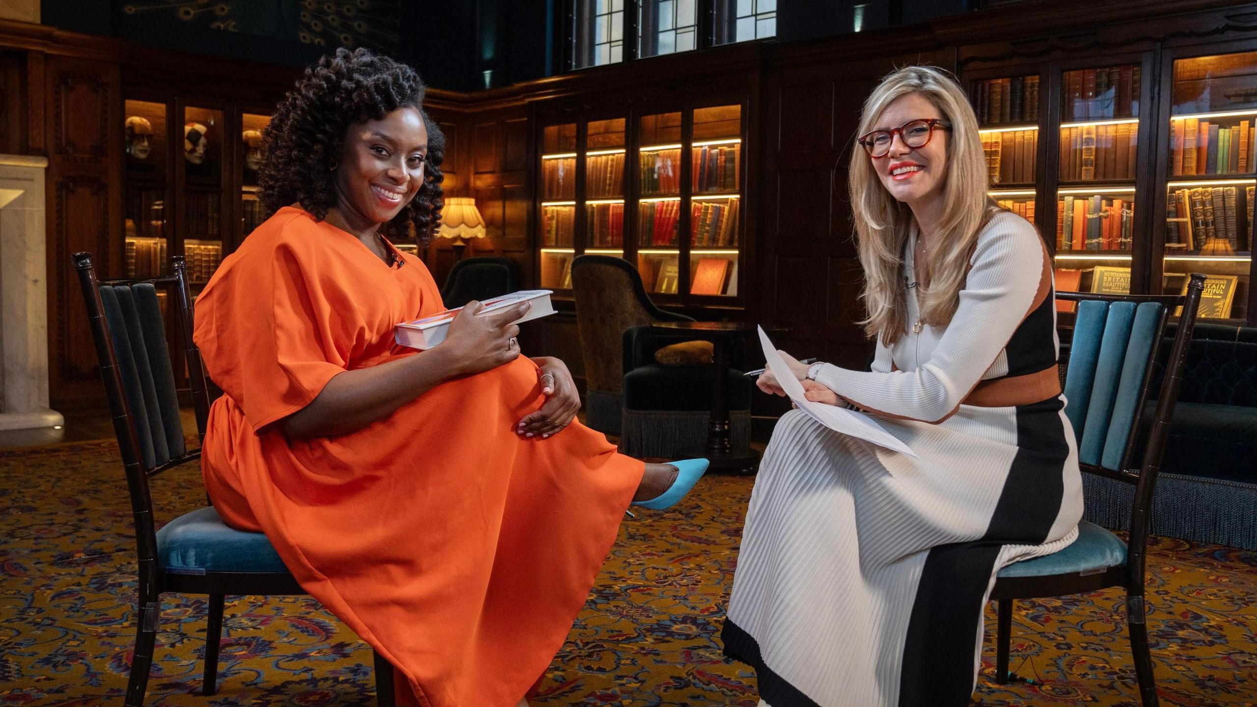 Chimamanda Ngozi Adichie and BBC journalist Emma Barnett sitting side by side. Ngozi Adichie is holding a book. Both are smiling. 