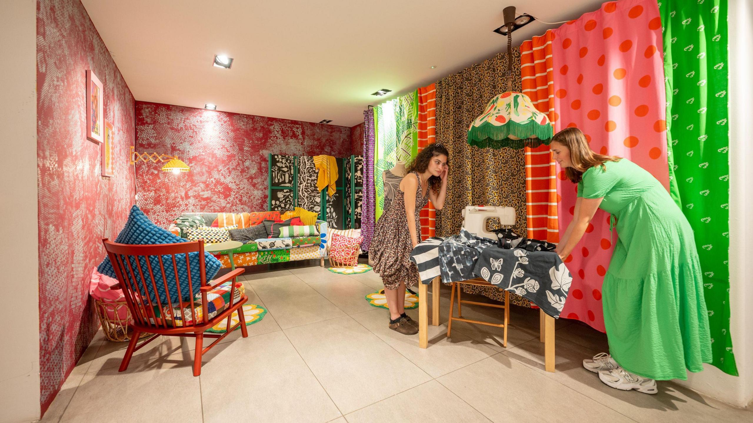 A room setting in the exhibition shows a colourful patchwork sofa with lots of different patterns, in a room with red Oriental-style floral wallpaper and a wall of curtain panels of very different bold patterns. Two women are lifting a sewing table into place.