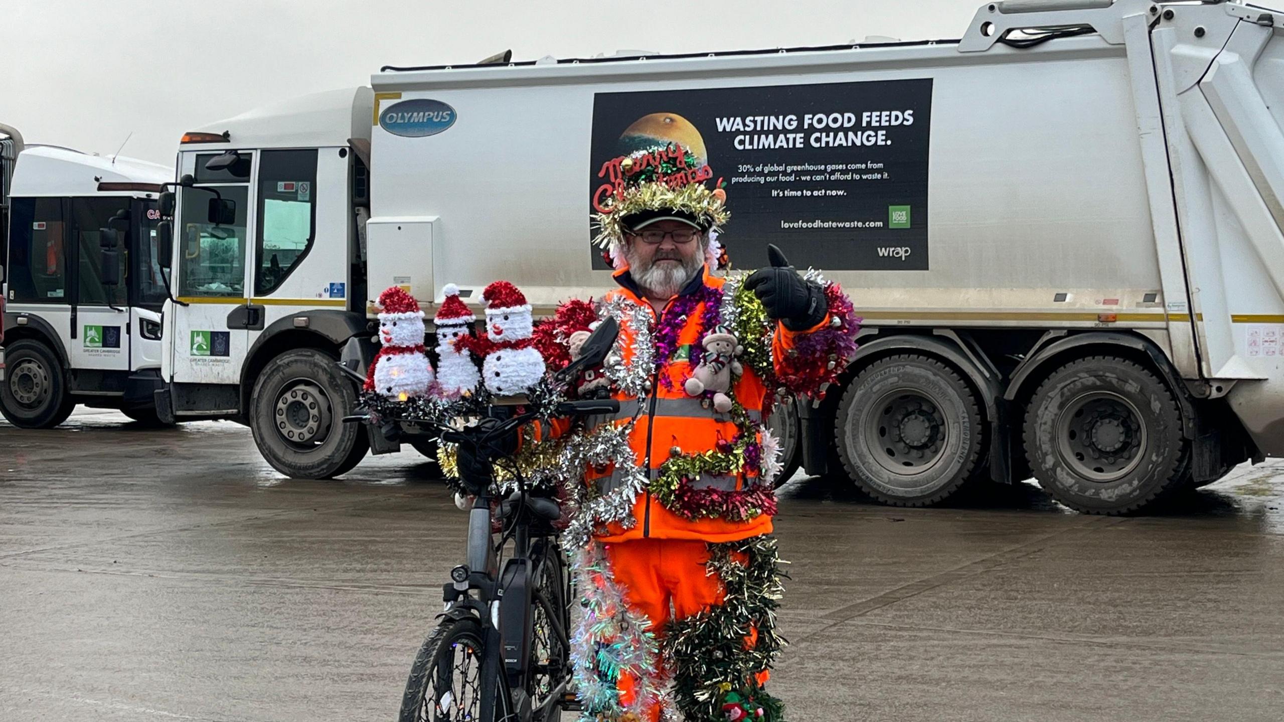 Lenny Edward stood in front of a bin lorry with a bike. He has decorated his bike with three white snowmen in red Santa hats. They are sat on top of his handlebars. 