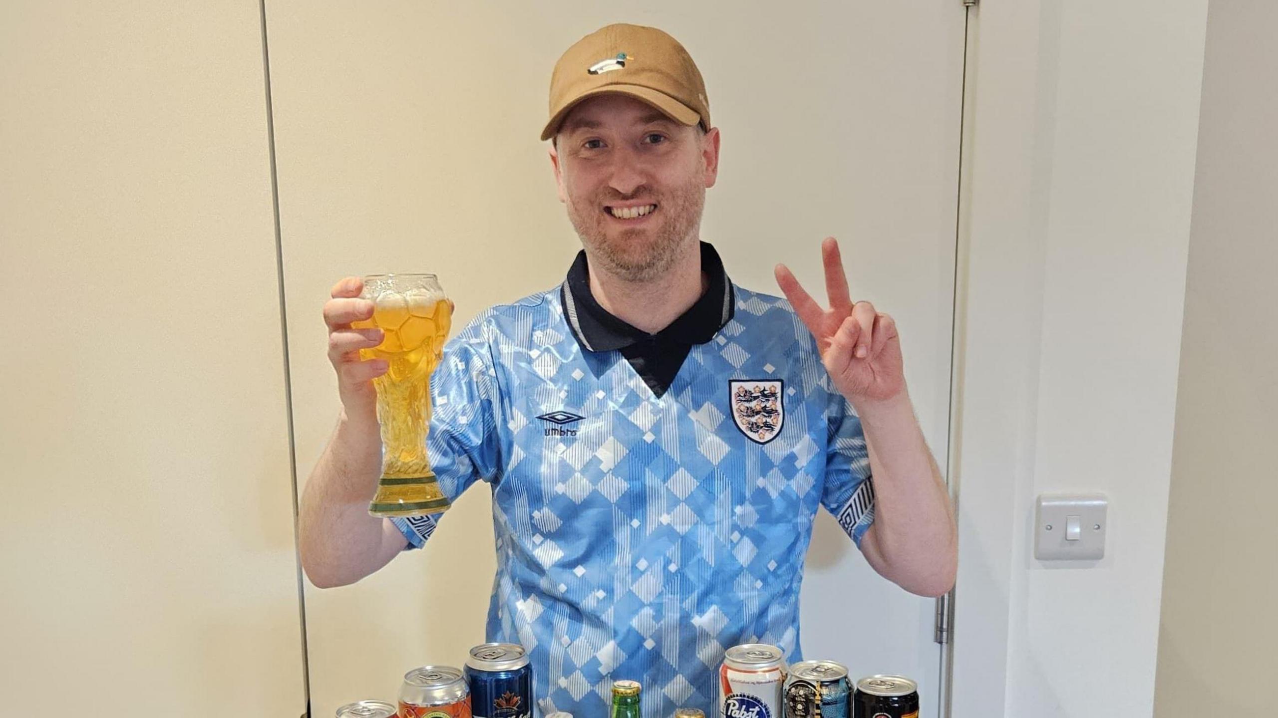 Gus Hully holds a beer in a glass shaped like the World Cup trophy, while he wears a vintage light blue England shirt and a brown baseball cap