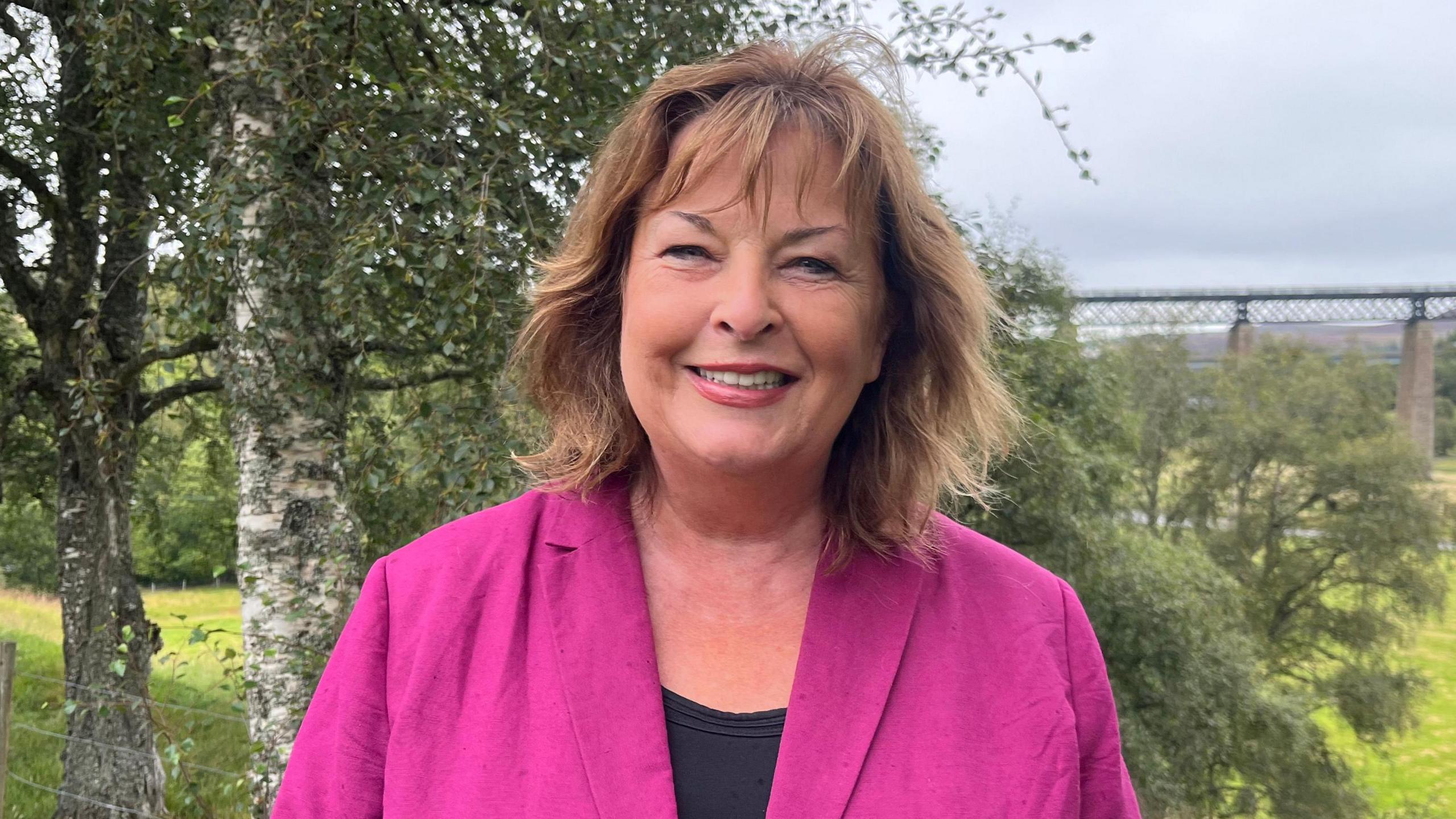 Fiona Hyslop standing outside and looking into the camera. She is wearing a black top with pink jacket and has auburn hair. Greenery is behind her. She is smiling.  