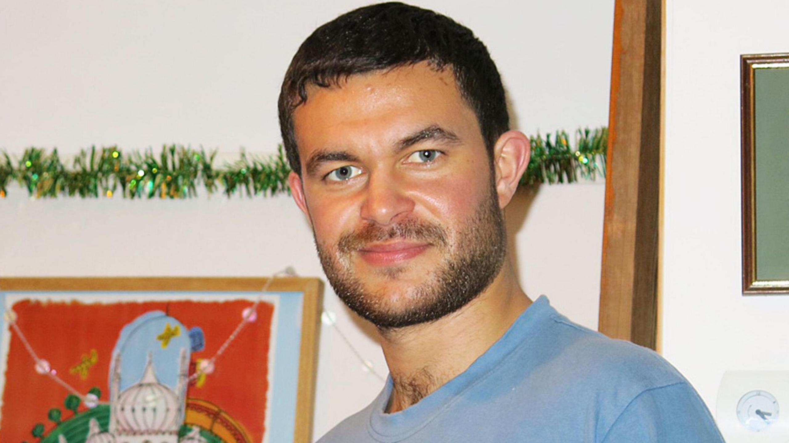 With short dark hair and beard, and wearing a light blue t-shirt, Matthew Grimstone smiles at the camera