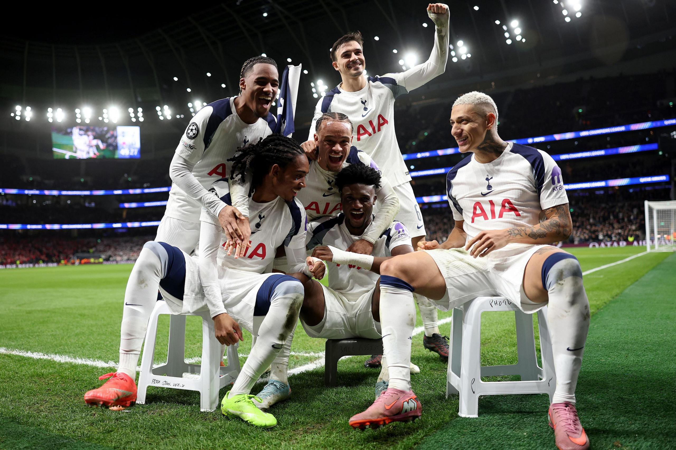 Un grupo de jugadores del Tottenham Hotspur, vestidos con camisetas blancas, se sientan juntos en pequeños taburetes al costado de una cancha de fútbol, celebrando y posando durante un partido dentro de su estadio brillantemente iluminado.