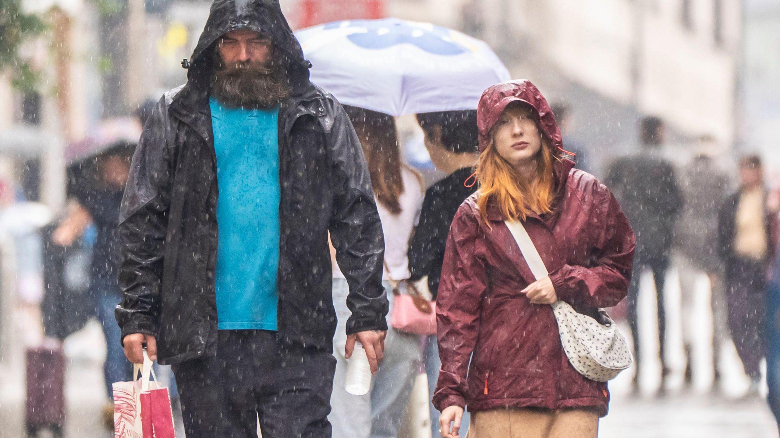 A man and woman walk along a street in Leeds, wearing rain jackets with hoods up as rain pours down.