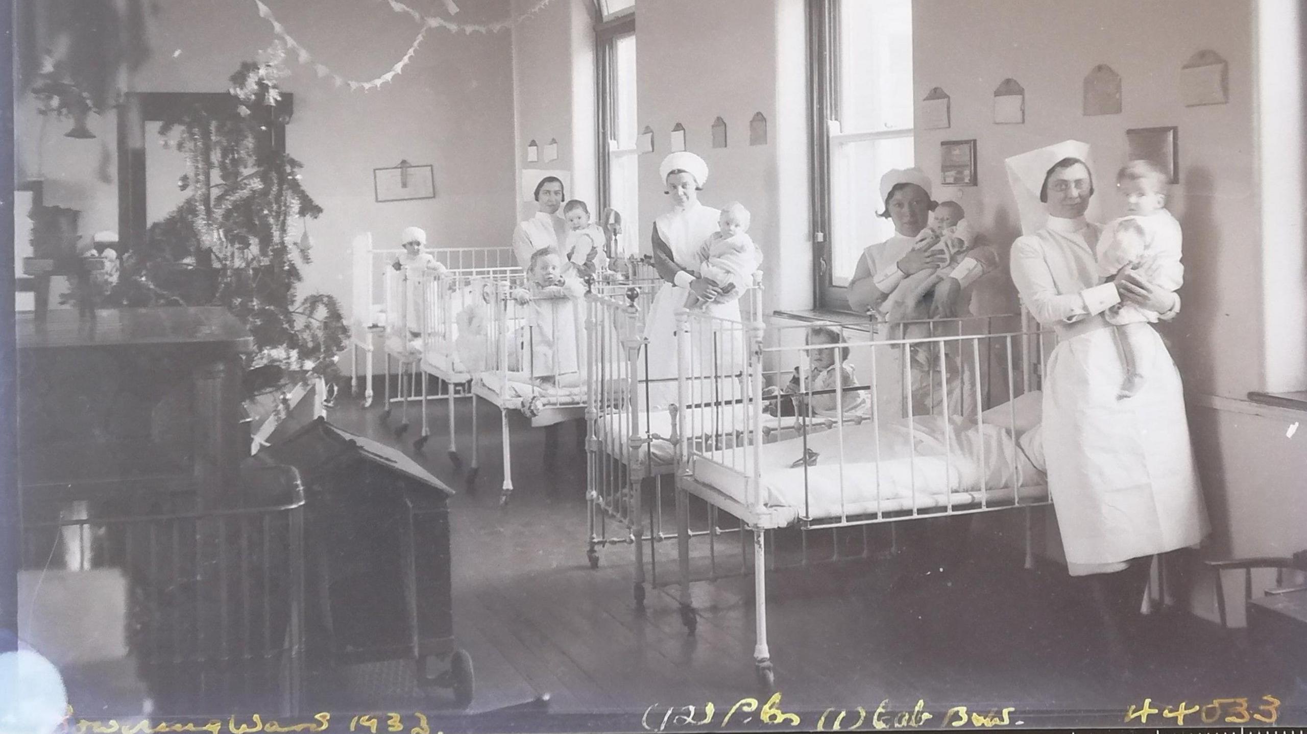 Nurses dressed in uniforms with aprons and headdresses hold babies beside their hospital beds in Bowring ward in the Royal Devon and Exeter Hospital. There is a Christmas tree in the background.

