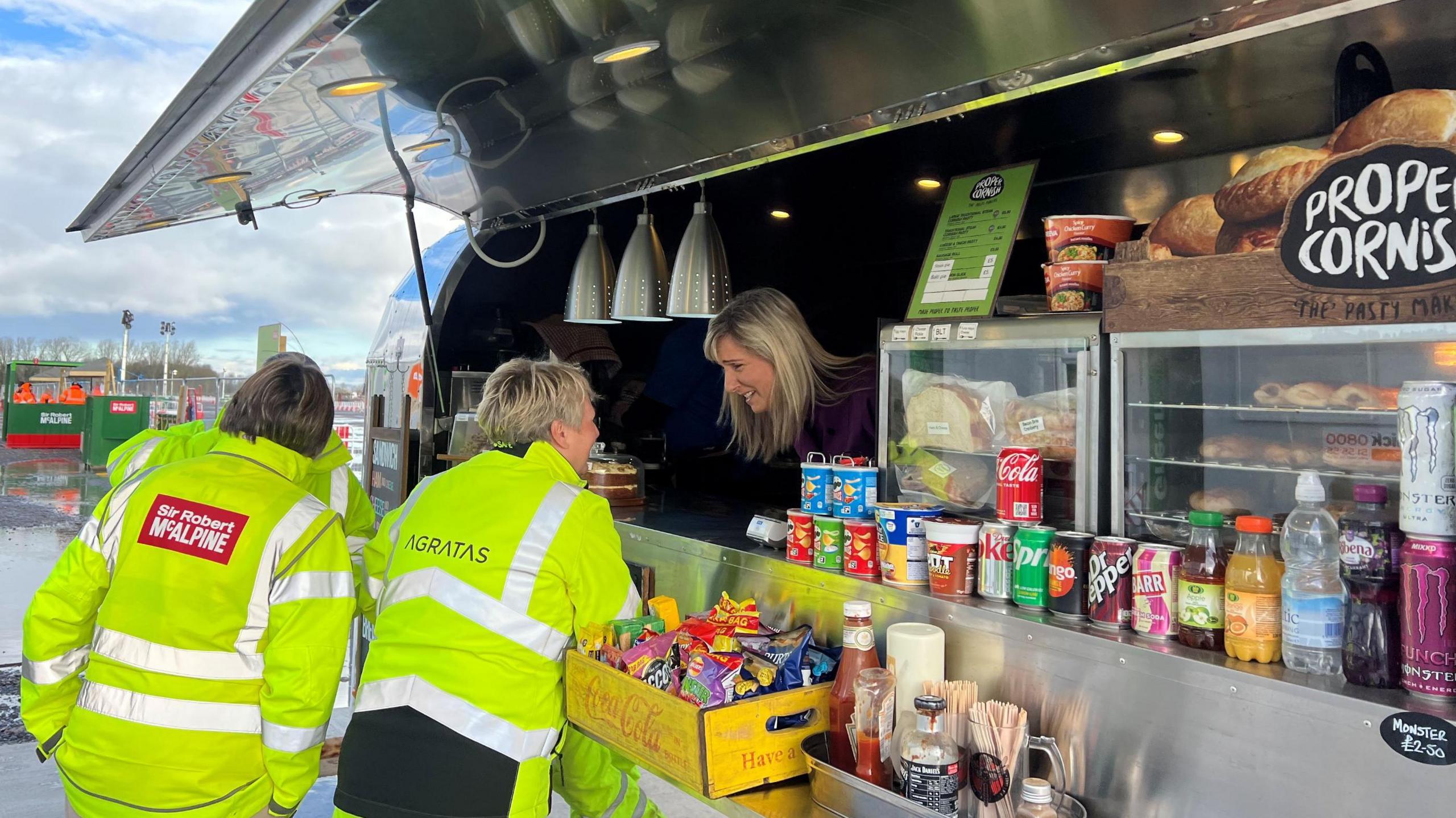 A woman leans forward out of a food van to serve two customers, who both have their backs to the camera and are wearing bright yellow high-visible yellow jackets. The food van is near the Hinkley Point C site in Somerset. It is made of light metal and various types of food and drink for sale are also visible