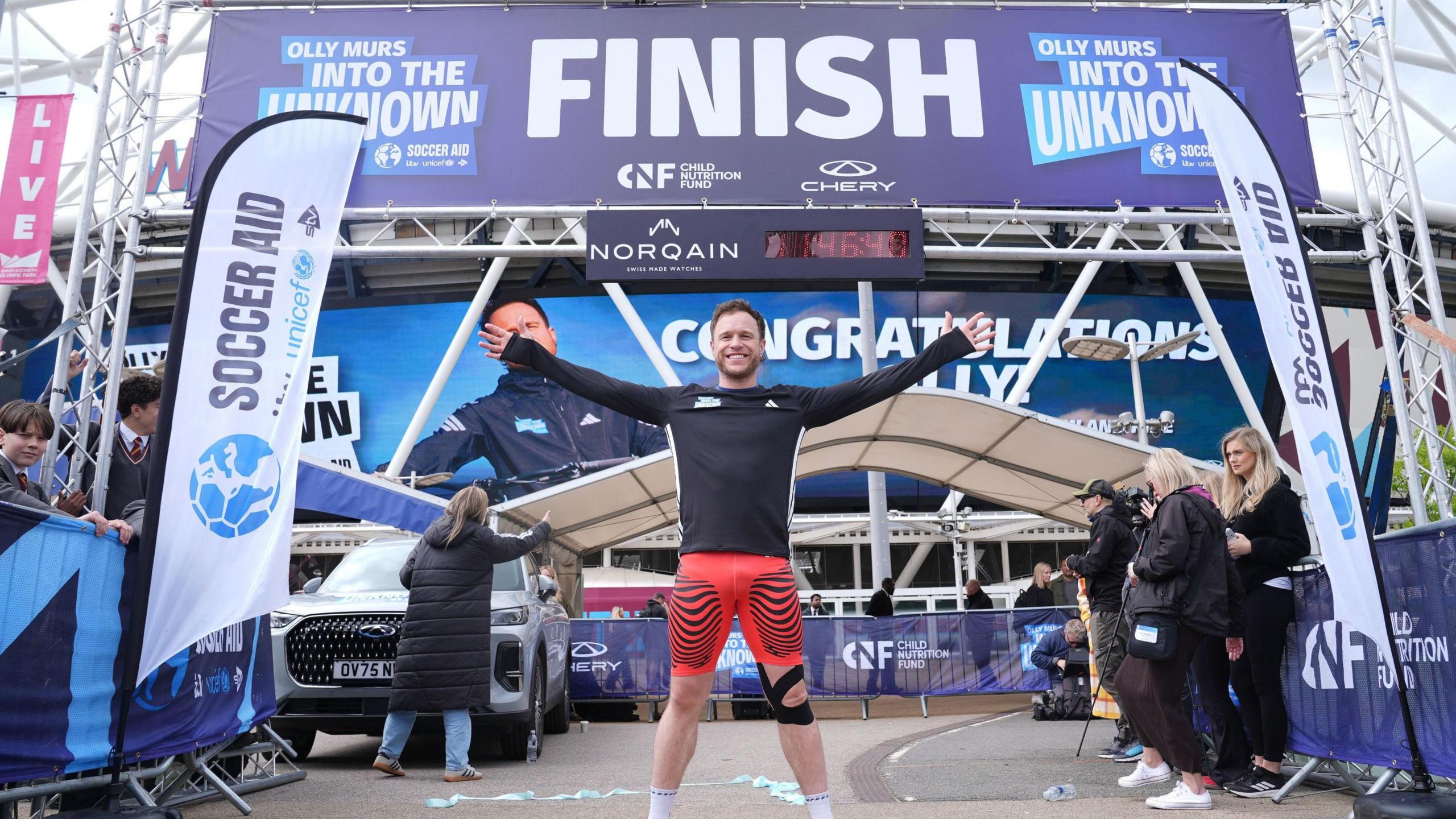 Singer Olly Murs crosses the finish line with his hands in the air wearing orange shorts and a black top