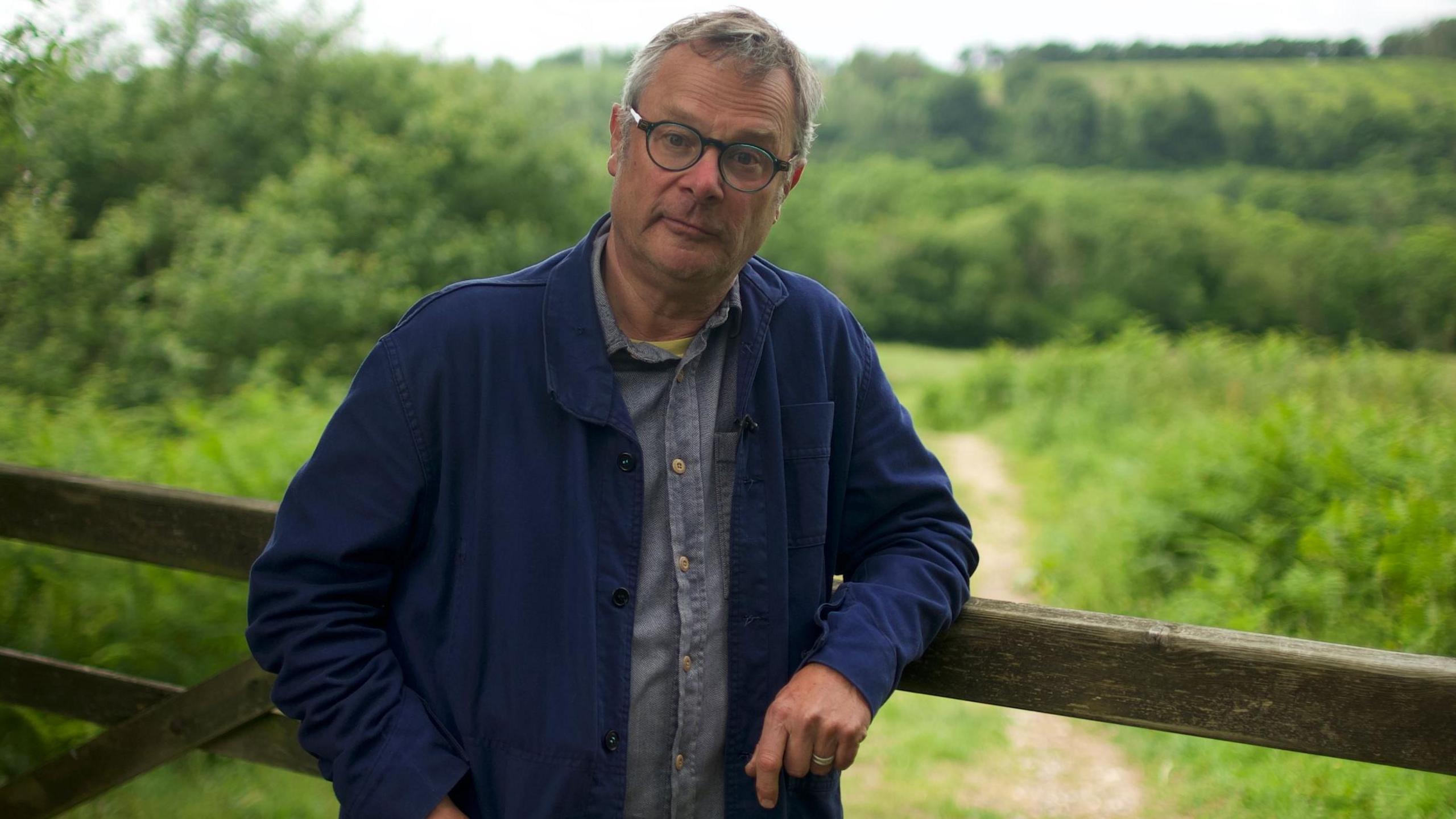 Hugh Fearnley-Whittingstall is leaning on a fence with green countryside behind him. 