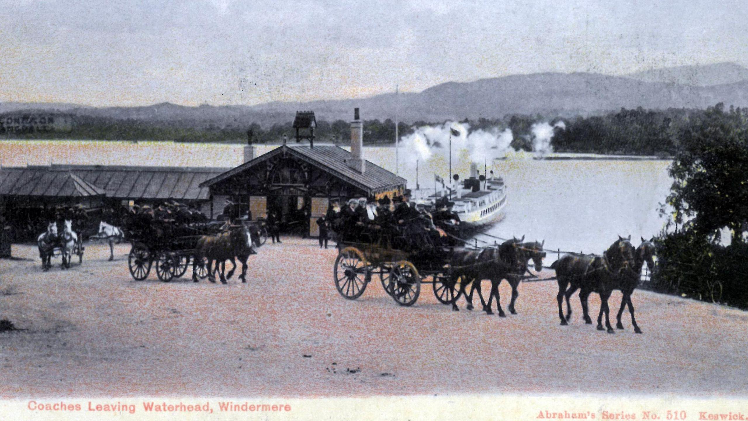 Historic photo in black and white, tinged with sepia and pale pink on the ground, showing horse-drawn open coaches, filled with passengers. Behind them, a steamer is drawn up next to a ferry landing on the shore of Windermere. Mountains can be seen in the distance.