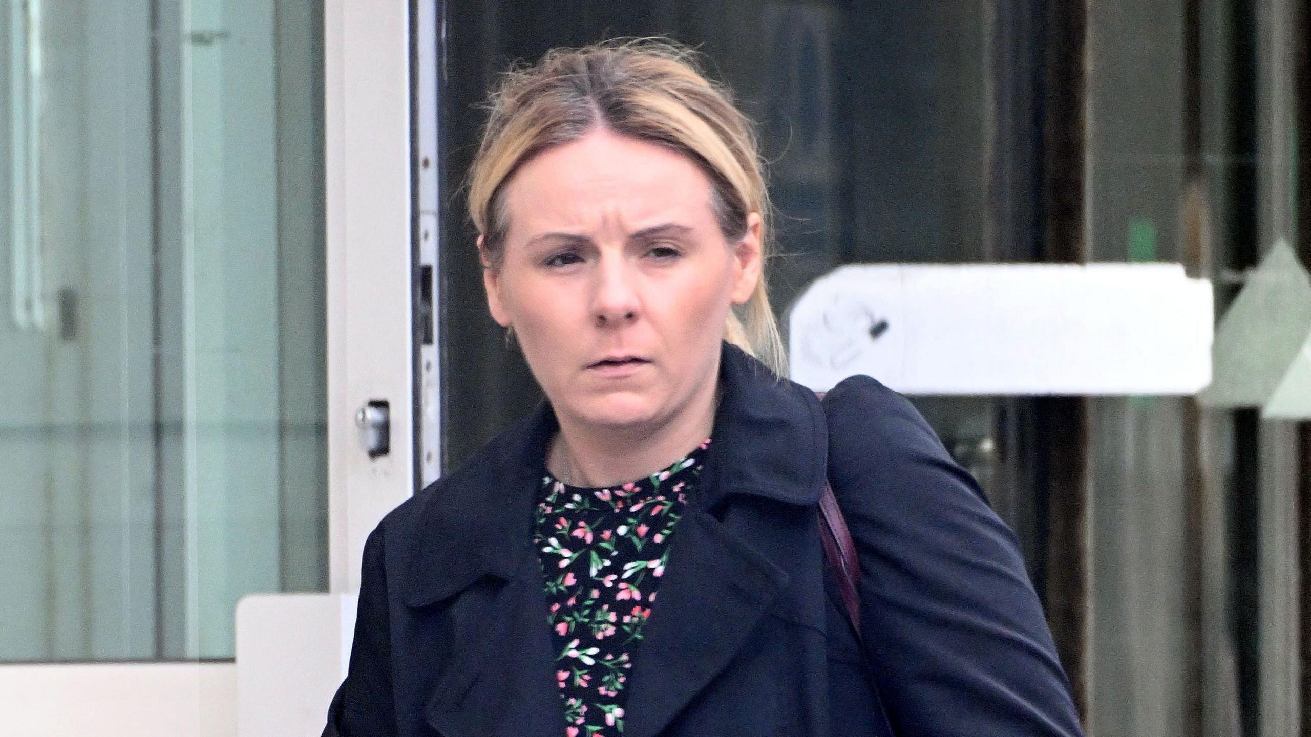 Judith Evans standing in front of a glass door. She is wearing a blue coat, a flowery top and carrying a handbag.