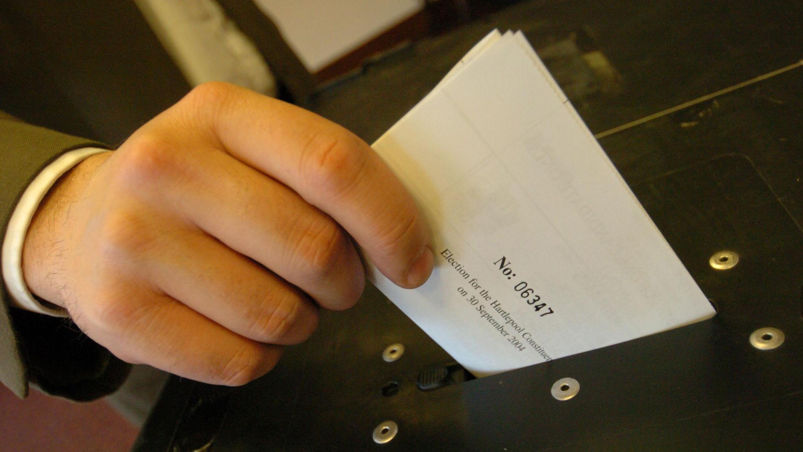 A hand, with a brown jacket visible on the sleeve, holds a folded white ballot paper in his thumb and forefinger. It is just above the slot on a black ballot box.