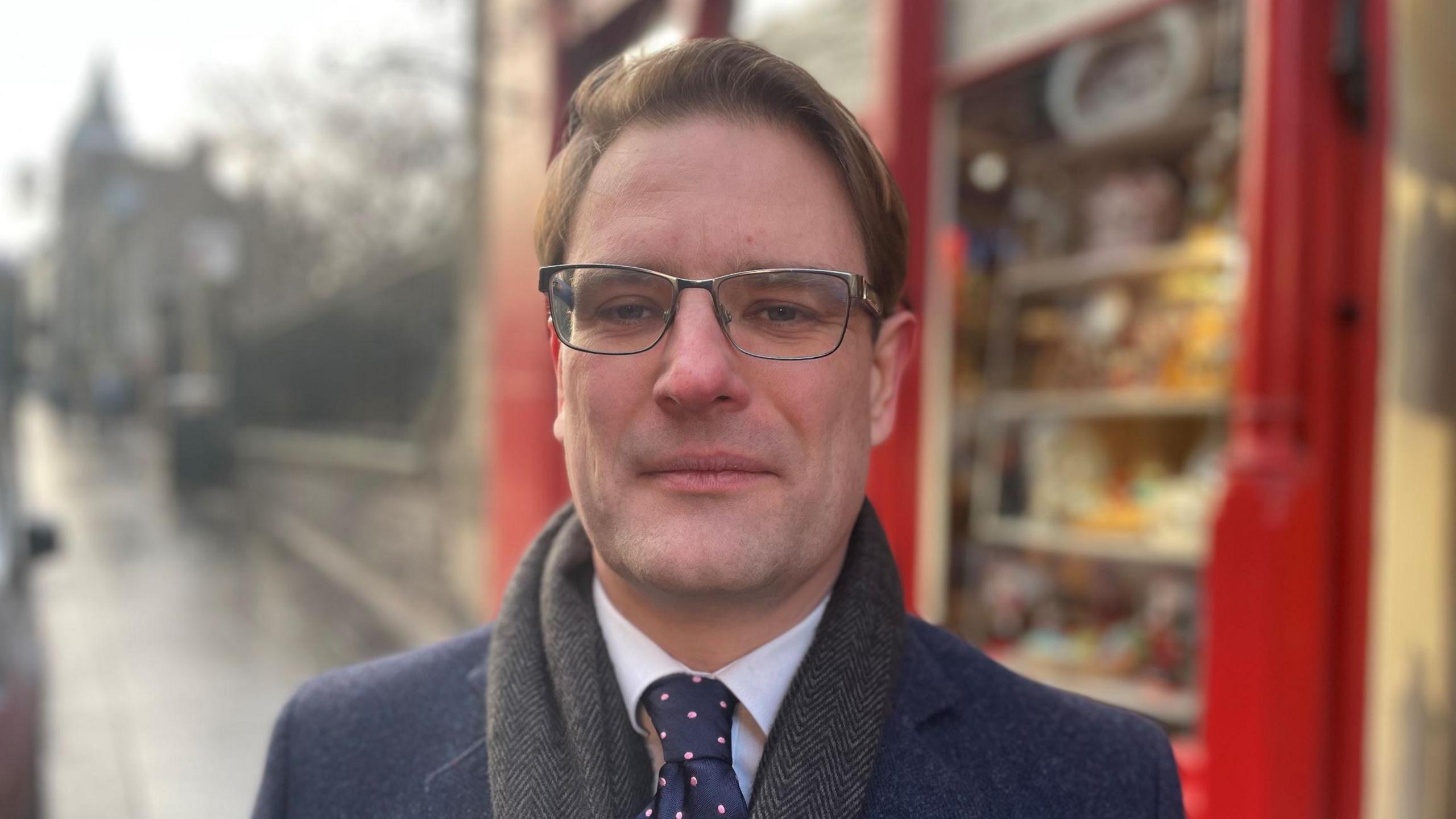 A head shot of Ewan MacDonald-Russell standing in a street, with a shop behind him. He has brown hair, glasses and a grey scarf on. He is looking directly into the camera. 