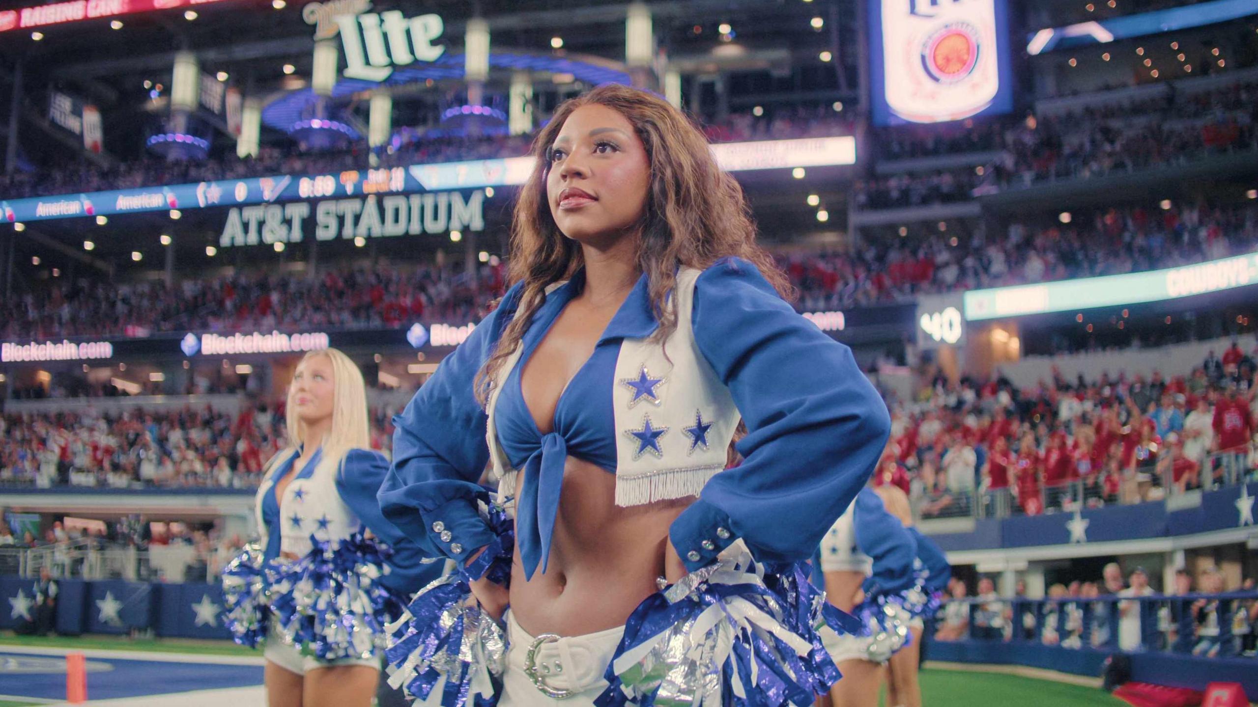 Two dancers on the field look straight ahead. The dancer in the front has long brown hair and the dancer in the back has long straight blonde hair. They wear the signature blue and white Dallas Cowboys Cheerleaders uniform 