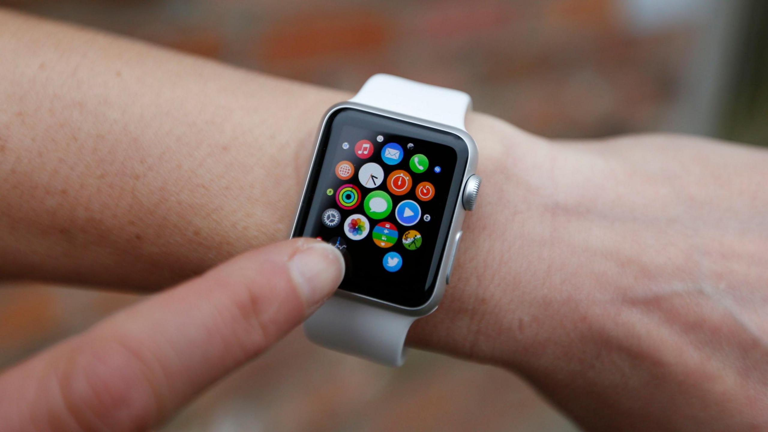 A smartwatch on someones wrist. There are multicoloured apps on the screen and a finger pointing towards them. 