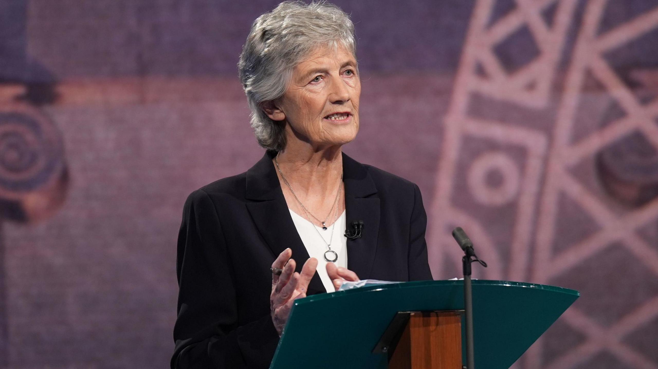 Catherine Connolly wearing a black suit and white top. She has grey hair. She is standing at a lecturn.