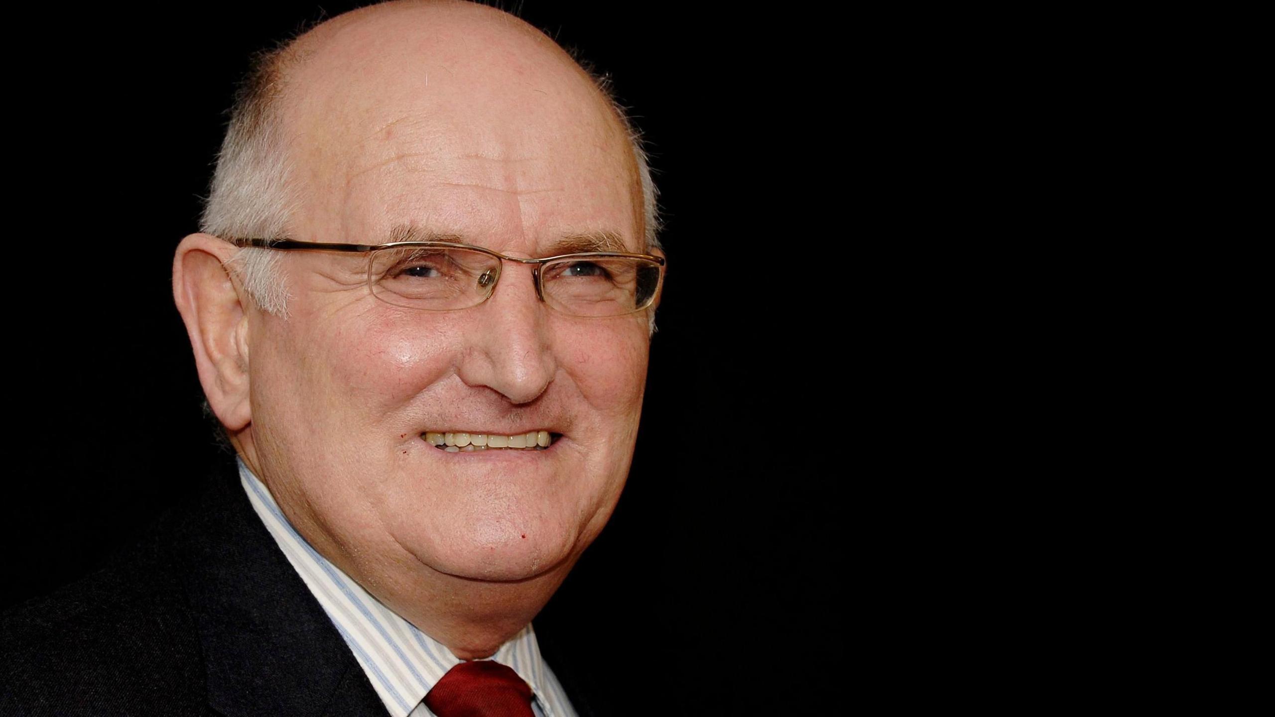 Head shot of Eddie Lowey wearing a red tie, light coloured striped shirt and dark coloured jacket set against a black background 