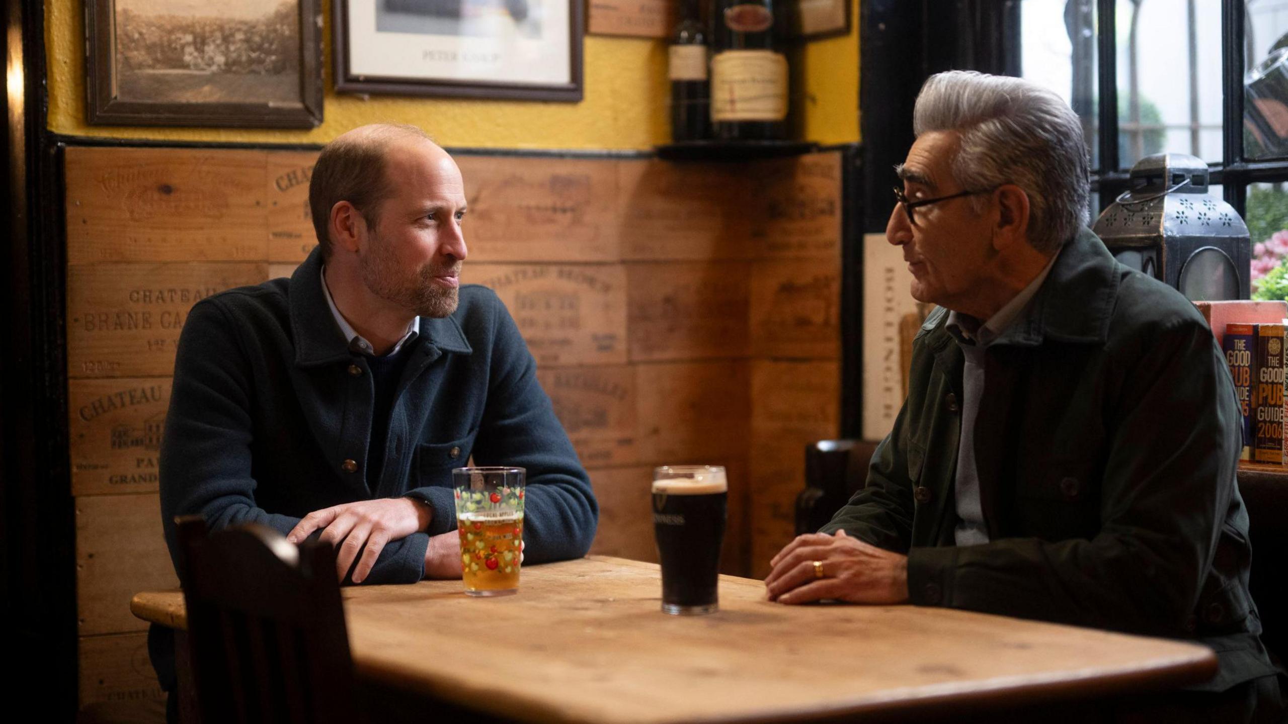 Prince William and Eugene Levy sitting in a pub with a pint each