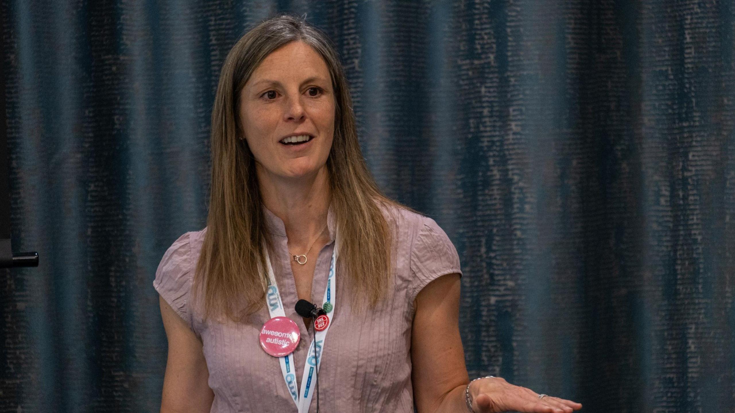 Kirstie speaks during a presentation. She has long light brown hair and wears a dark pink top. A blue curtain is behind her.