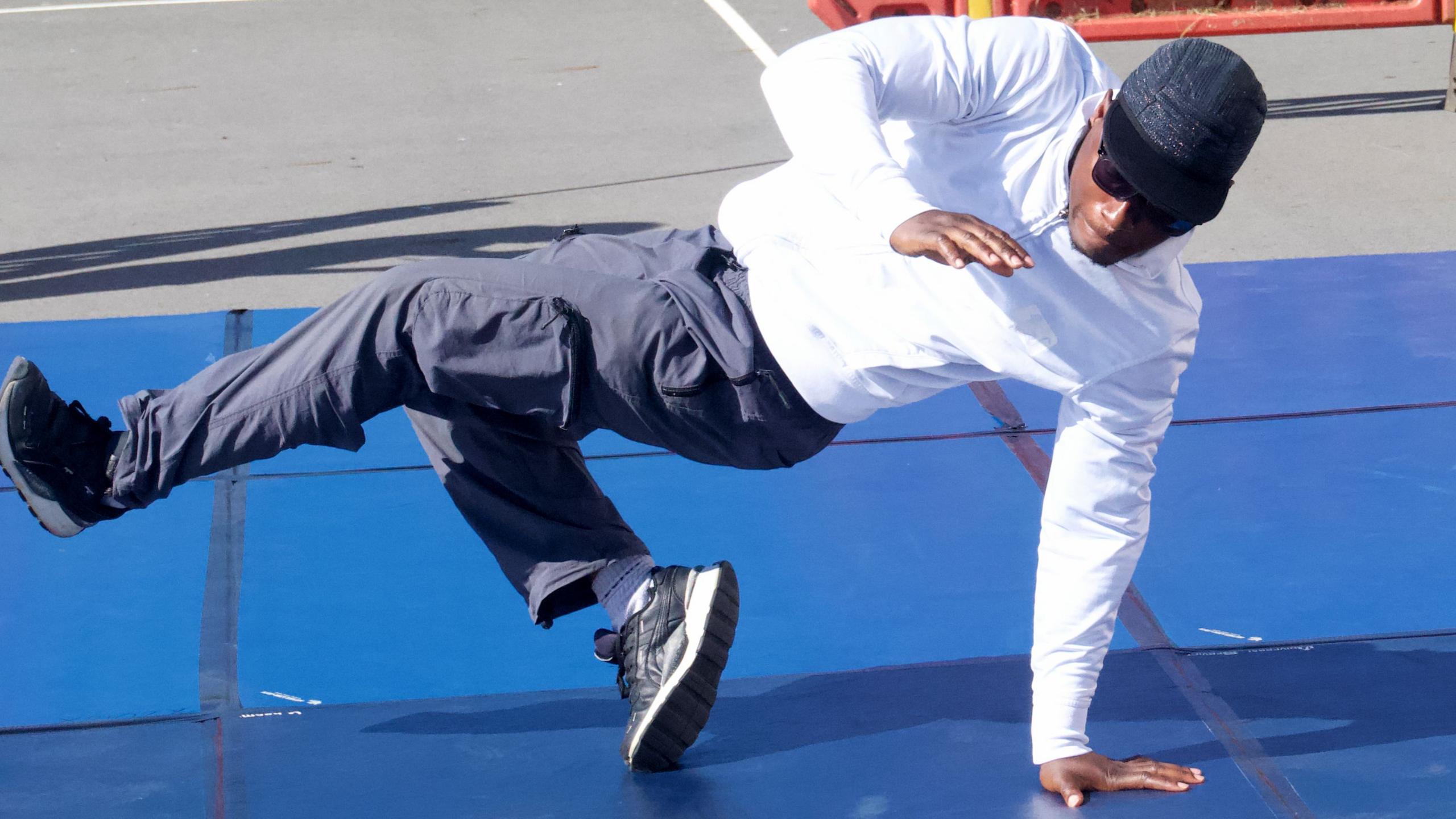 A man in a white jacket and grey trousers and a baseball cap performs a breakdance routine on a blue mat