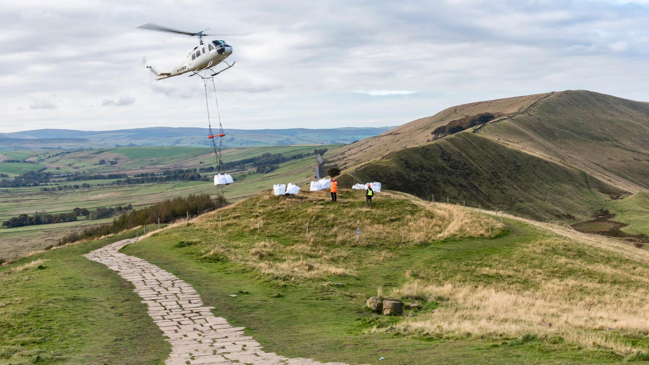 A helicopter carrying large white bags of stones over a grassy part of the countryside