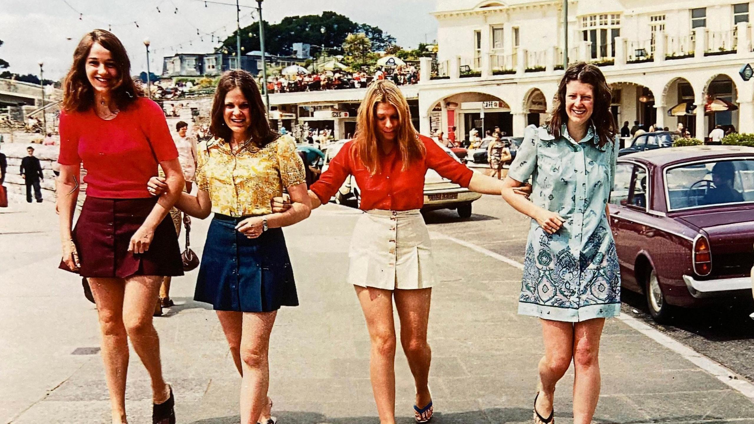 The same four women link arms on a seaside promenade in 1972. All are wearing bright, distinctive clothes in line with the fashion of the times. A white hotel is in the background.