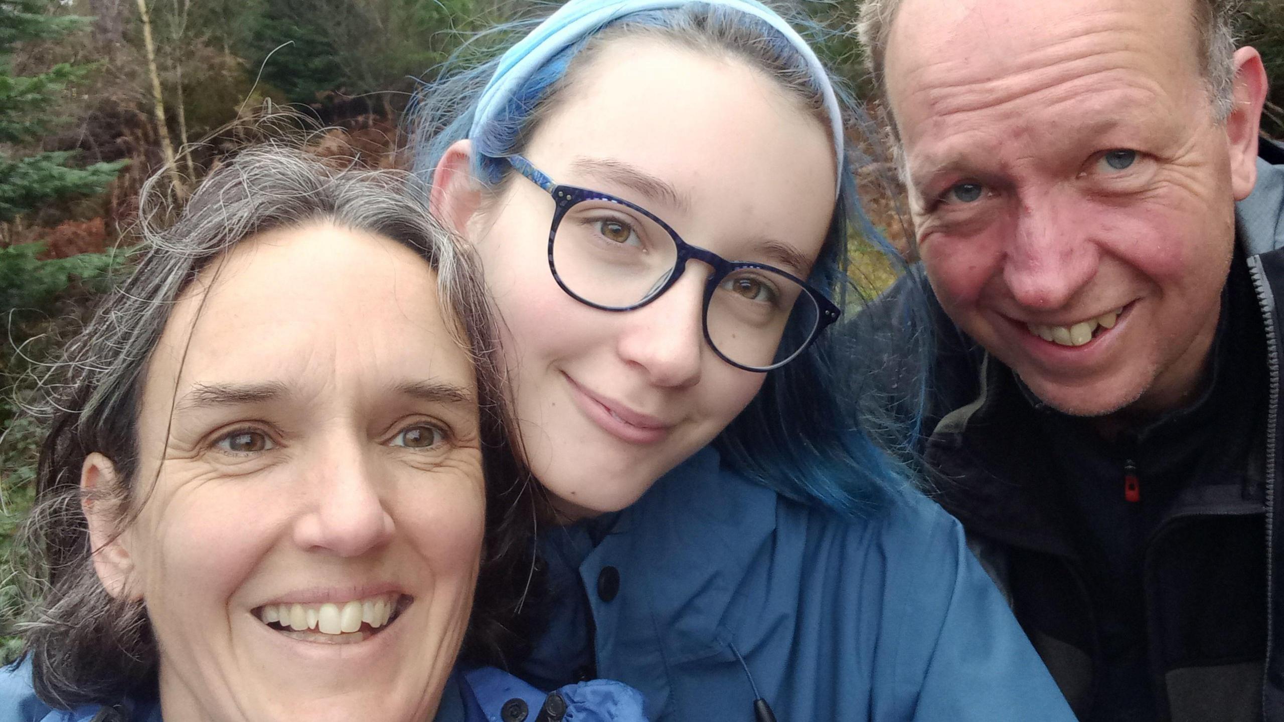 Katie Nellist, wearing glasses and with blue hair, stands with her arms around her mum, who is standing to her right. Katie's dad stands to her left. They are all smiling at the camera.