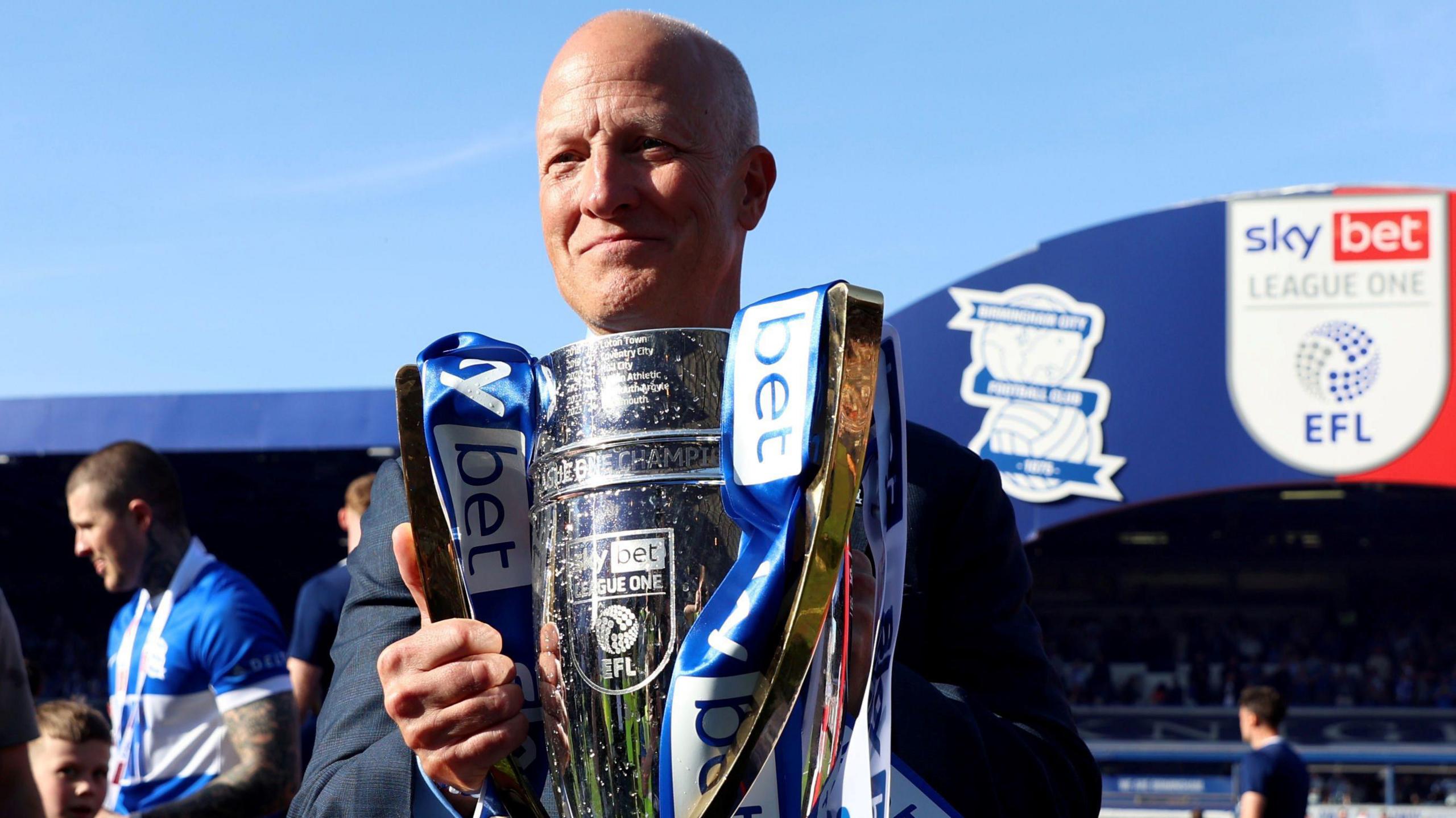 Birmingham City chairman Tom Wagner with the Sky Bet League One trophy after the Sky Bet League One match at St. Andrew's @ Knighthead Park, Birmingham. He is holding the silver trophy which has blue and white ribbons on it. Behind him is the current stadium and several fans. 