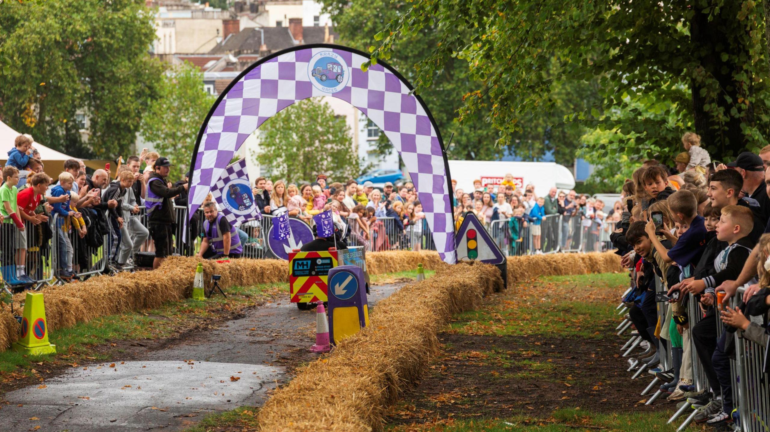 A solitary home-made cart, in the shape of a tube, with two people in it, races away from the camera as part of the Krazy Races event in Victoria Park in Bristol. Dozens of people can be seen either side of the tarmac track, which is bordered with bales of straw. There are also metal railings keeping the crowd back.