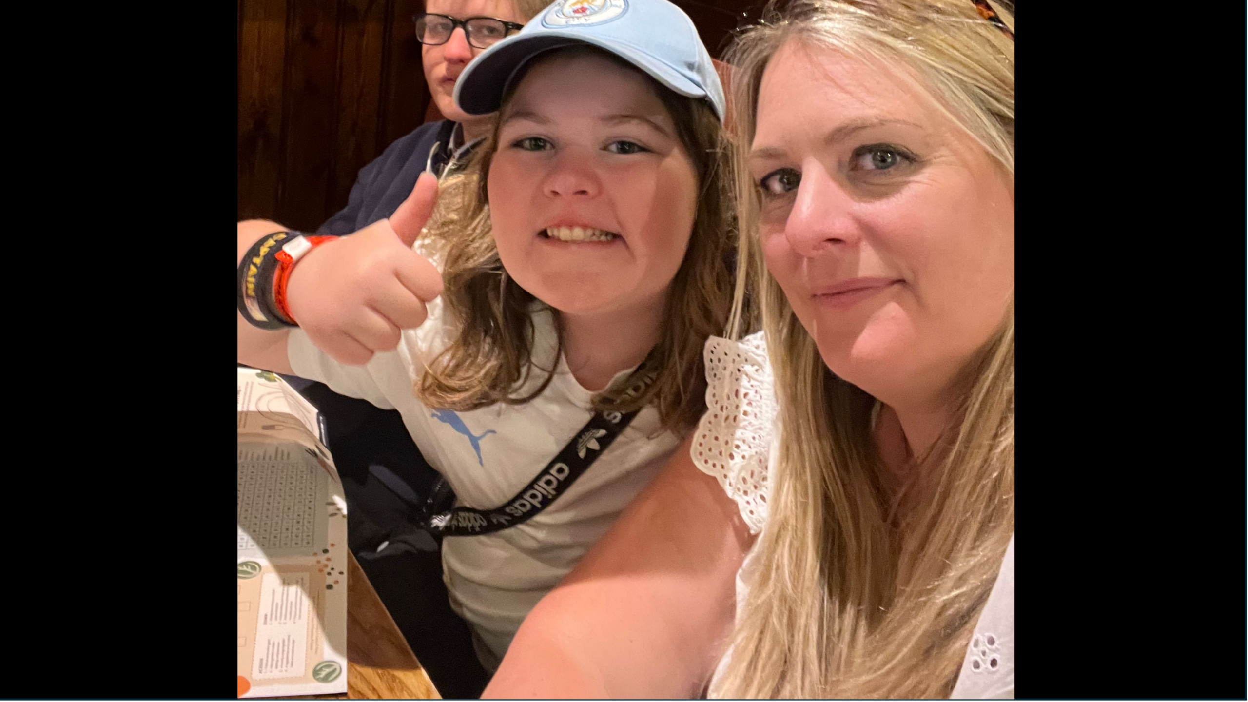 A girl with brown hair smiles and a blue cap smiles and gives a thumbs up to the camera while a blonde woman next to her takes a selfie with her