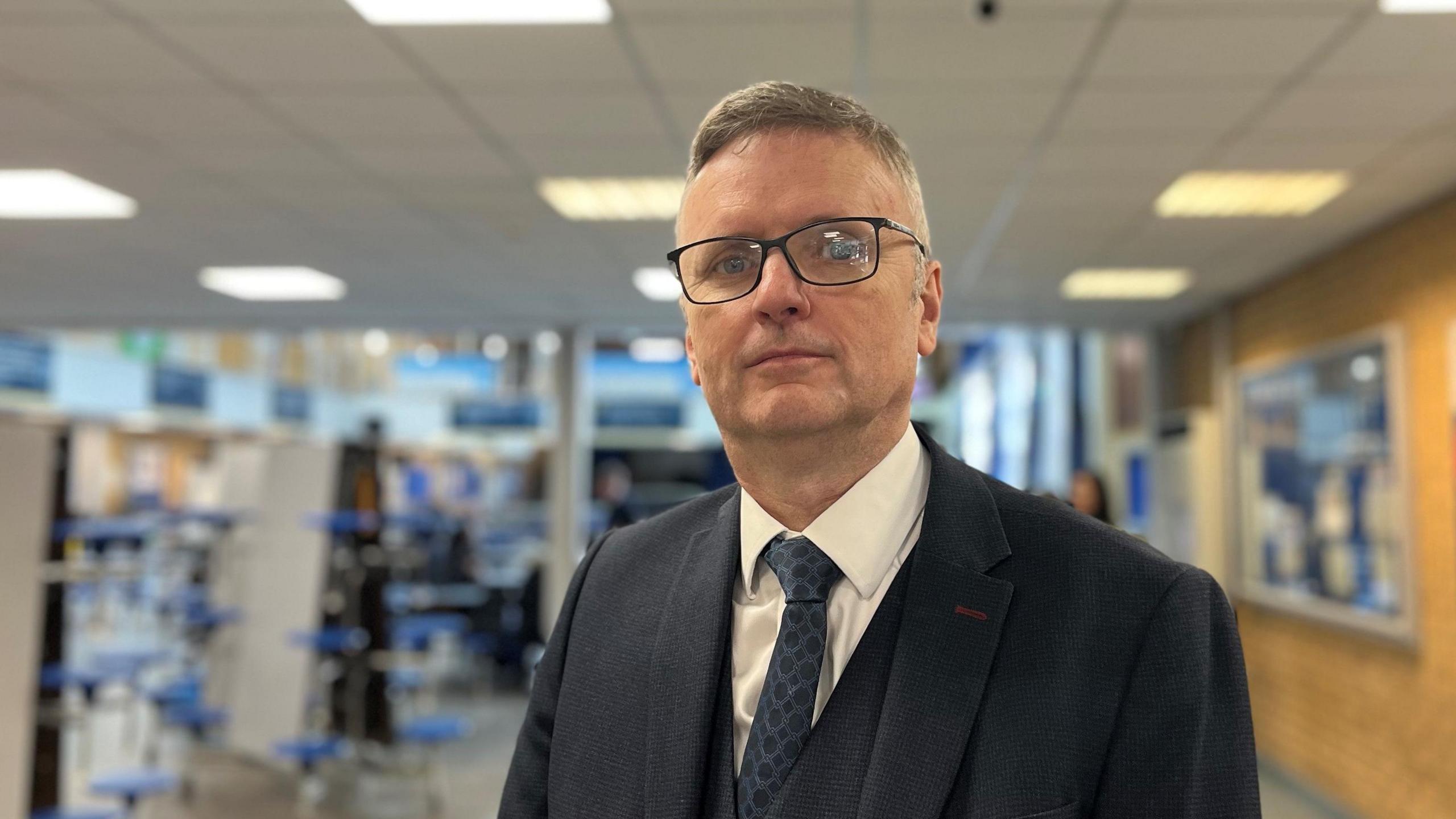 John McGhee standing in a school corridor looking directly at the camera. He is wearing glasses, and dark blue three-piece suit and a blue tie, with a white shirt.