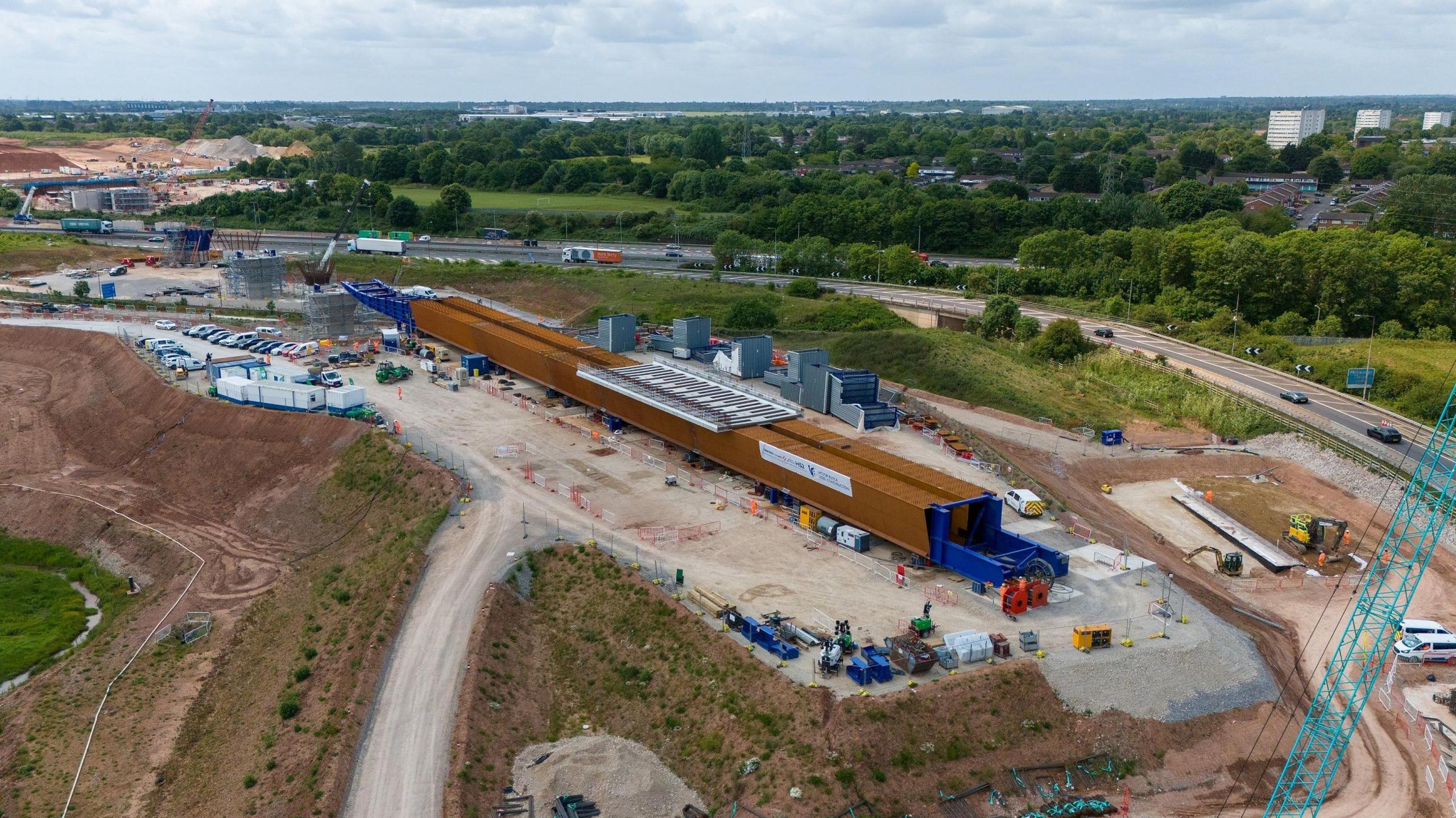 An aerial shot of a construction site next to a motorway, with green spaces and trees on the other side of the carriageway. A long brown structure is being built a few dozen feet from the motorway.