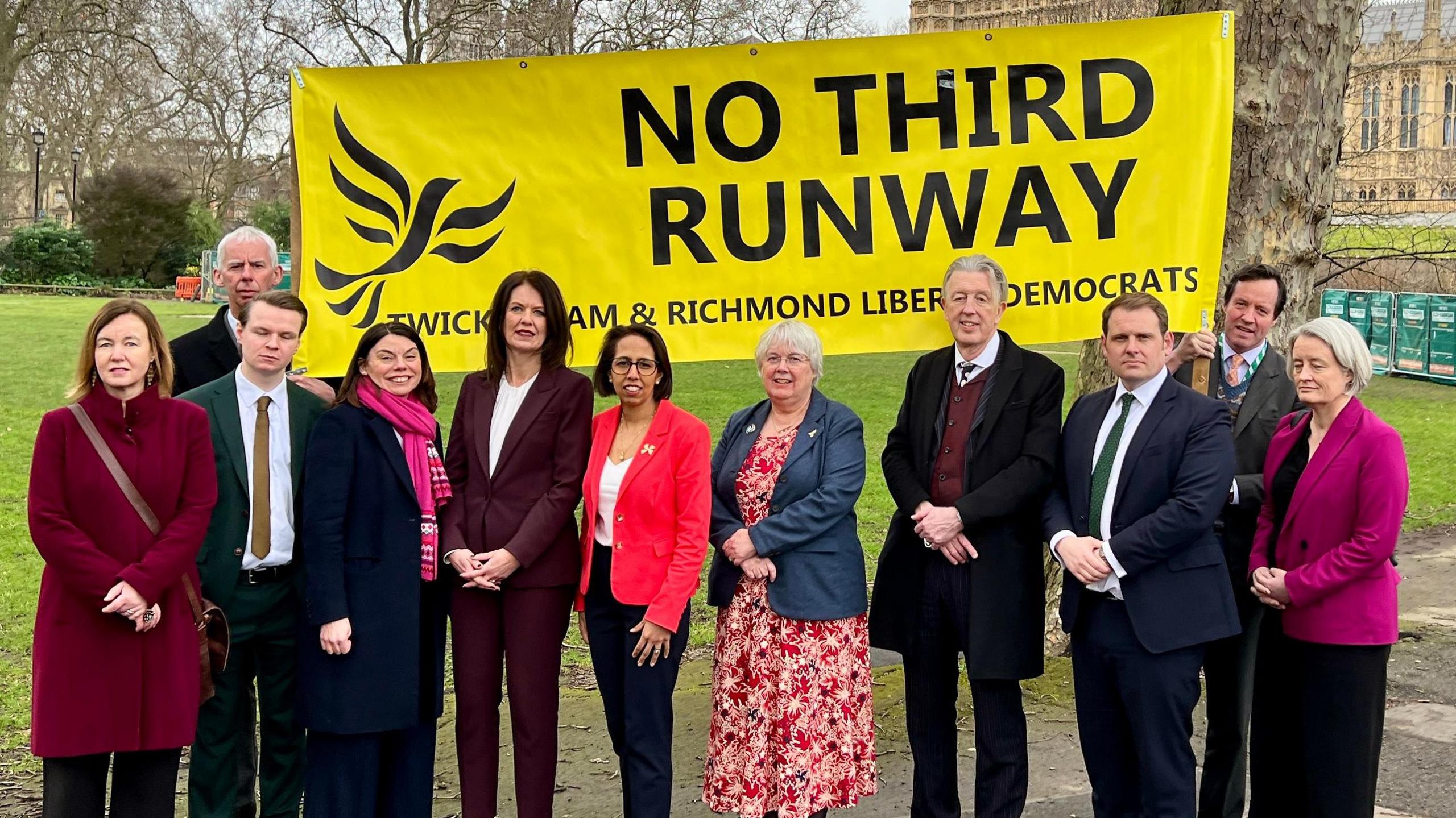 Eleven men and women stand in front of a large yellow banner which says No Third Runway with a Liberal Democrat logo. They are stood in a park next to the Palace of Westminster.