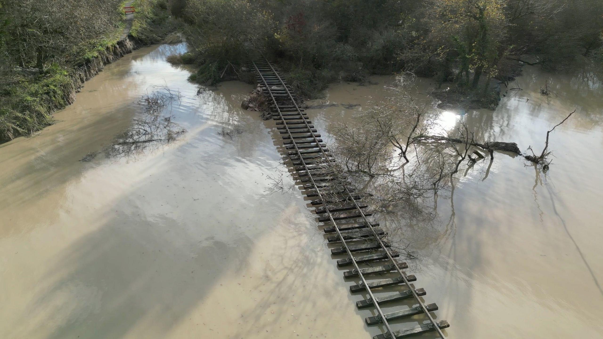 A rail line surrounded by brown flood water. There is a fallen tree to the right of the rail line. The line is having off the ended of the bank. 