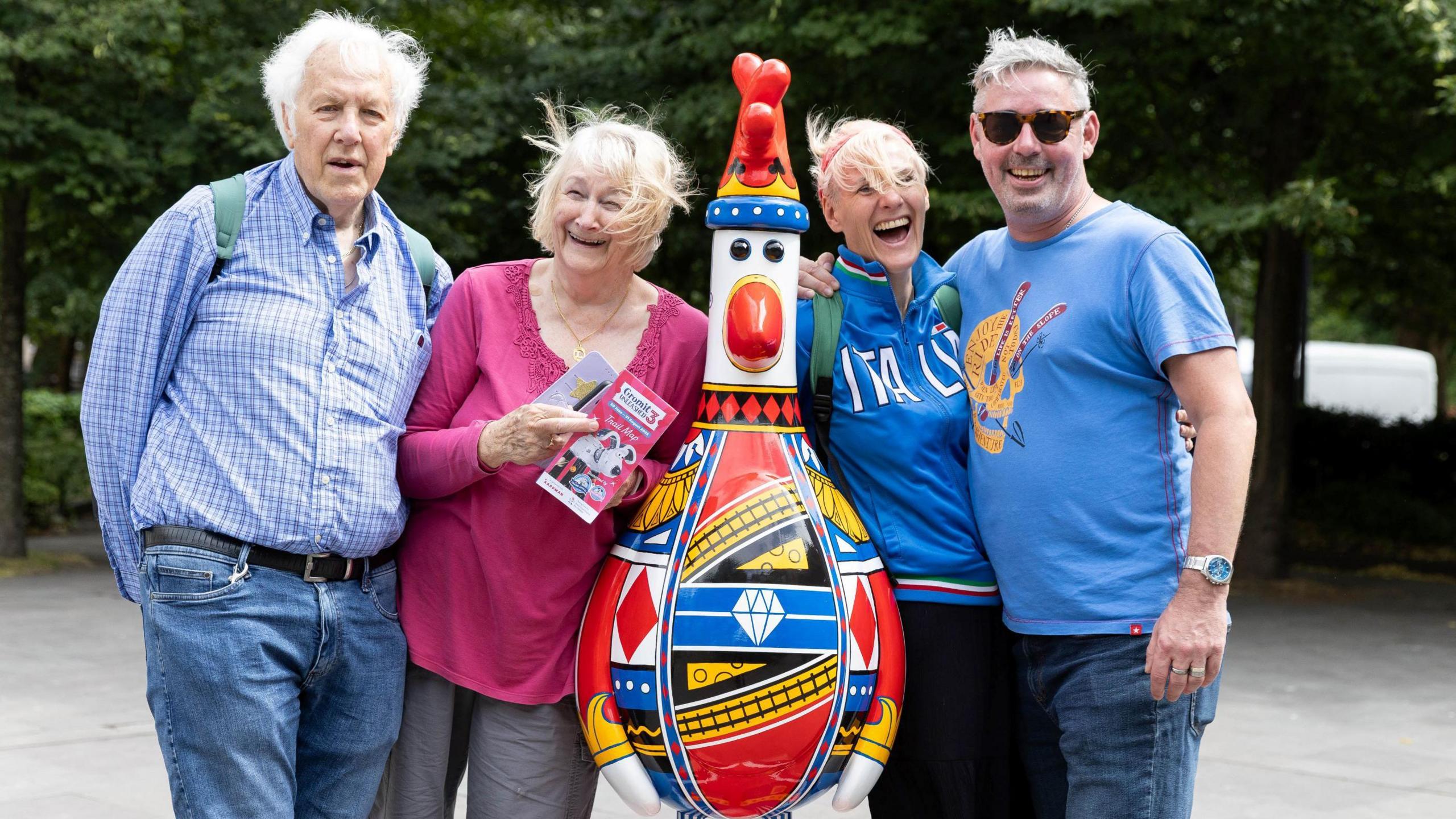 Four people, two men and two women, stand either side of a colourful Feathers McGraw statue, which is part of the Gromit Unleashed 3 trail in Bristol. They are all smiling at the camera and the statue is designed to look like the playing card the King of Diamonds.