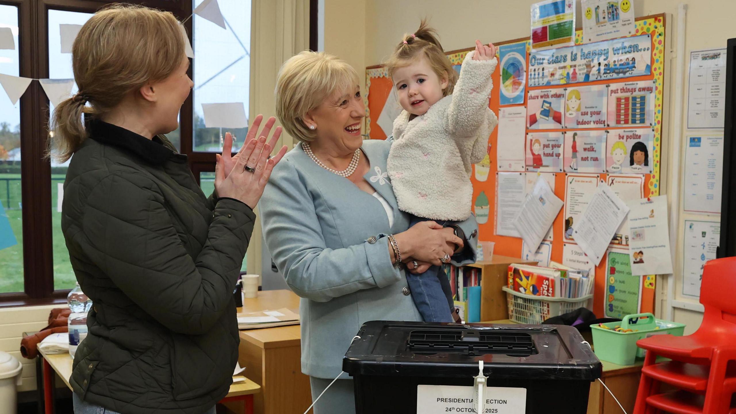 A woman in a light blue blazer and trousers stands in the centre of the image. She is holding a young child. On her left is a woman wearing a green jacket who appears to be clapping her hands. They are standing in front of a black box.