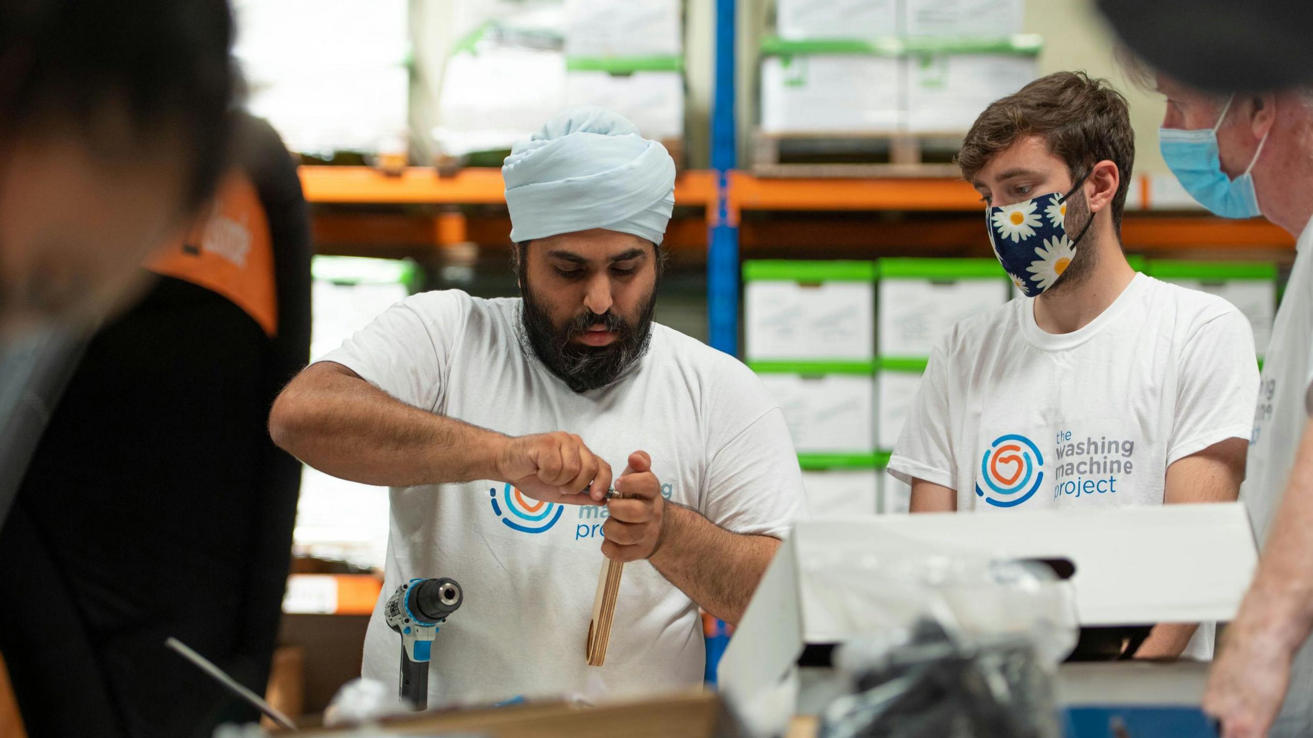 Founder Navjot Sawhney pictured building a washing machine in a warehouse, alongside other people also wearing Washing Machine Project t-shirts.