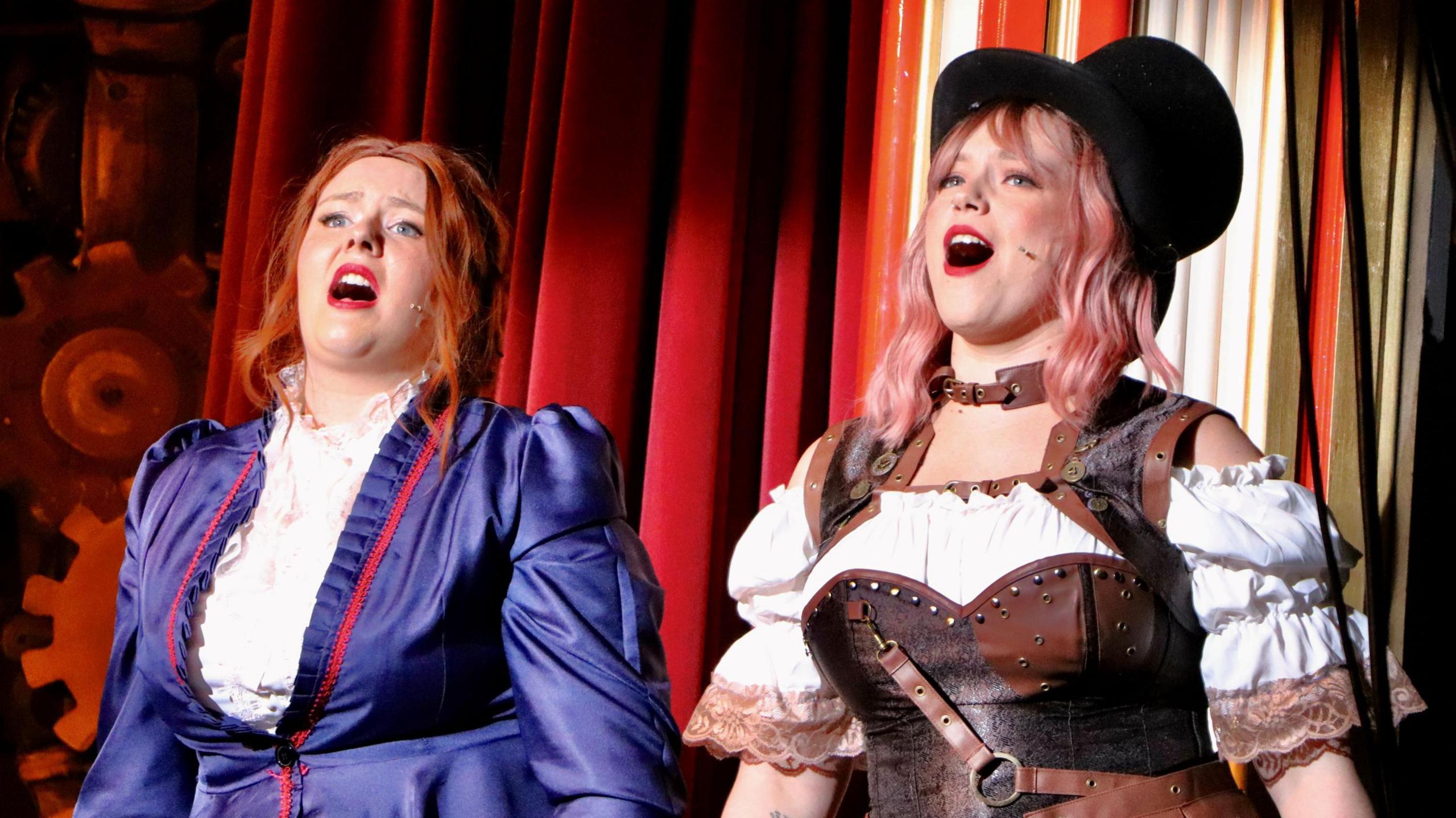 Two women in period costumers, one with a top hat, singing on stage. There is a red curtain behind them.