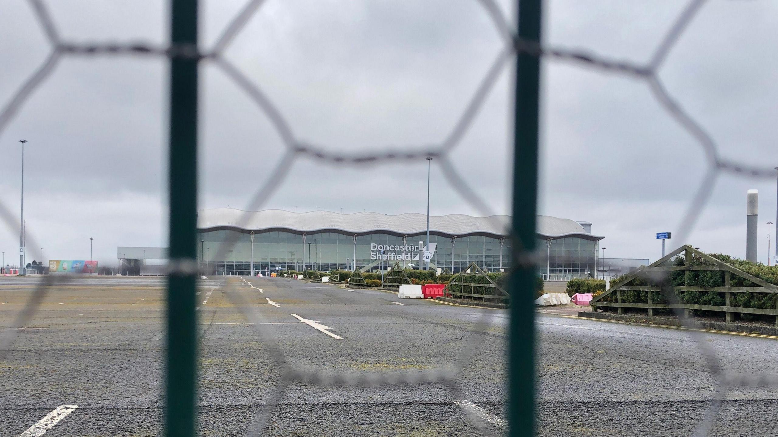 Doncaster Sheffield Airport terminal building surrounded by empty carparks. The picture is taken through a wire fence. 