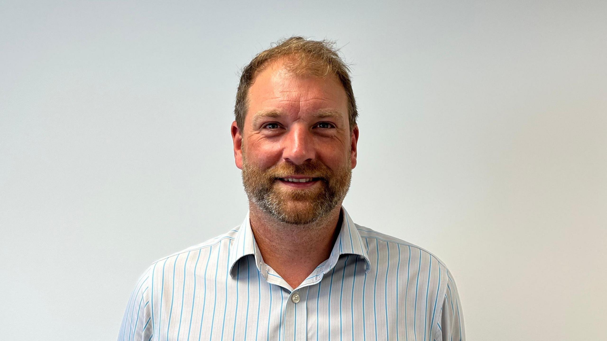 Jamie Baldwin, a man with light brown hair and stubble, he smiles and wears a white stripy shirt in front of a white background.