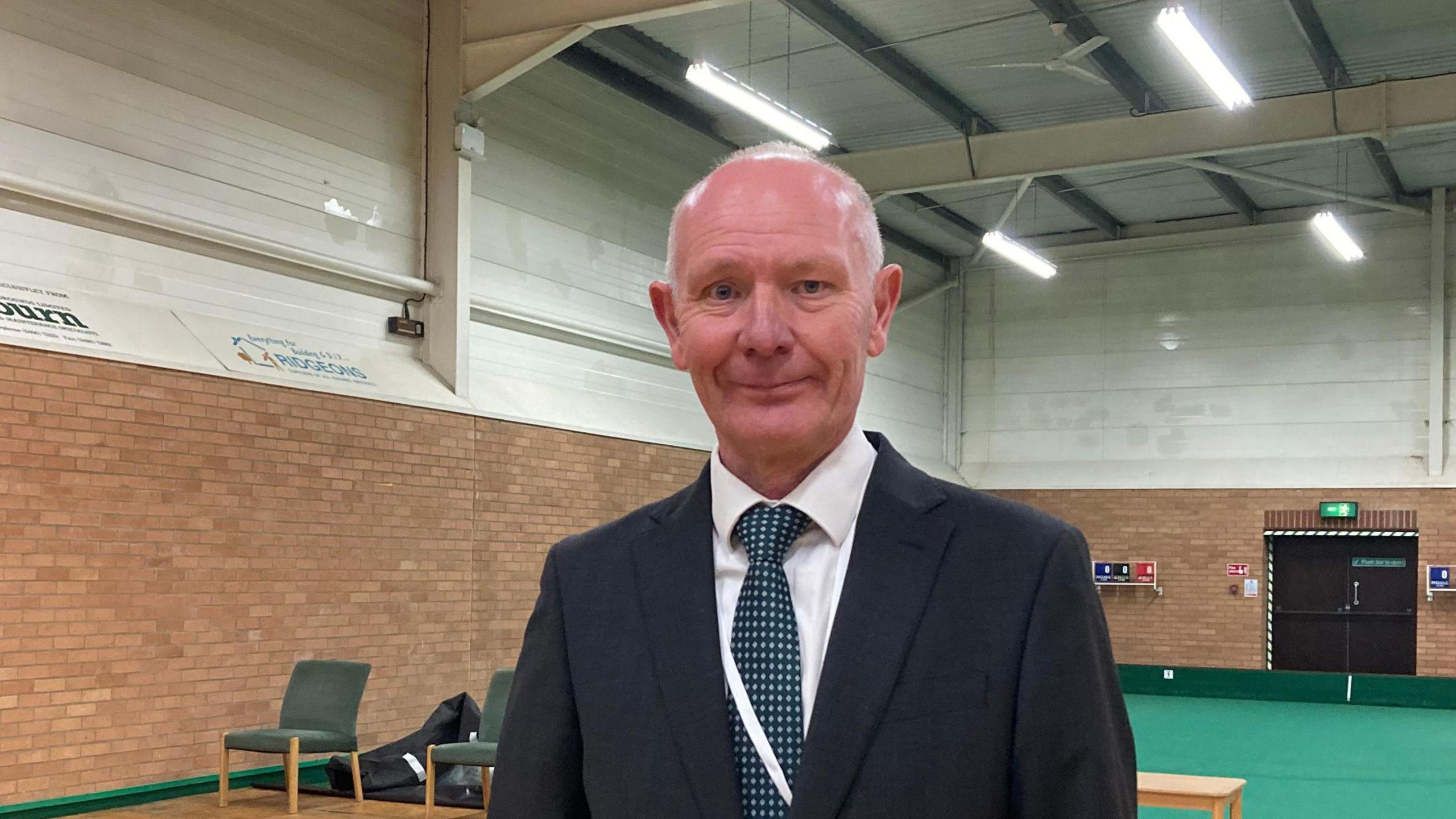 Darryl, a man wearing a black suit and white shirt with a printed black and white tie, smiling for camera, standing inside a sports hall.