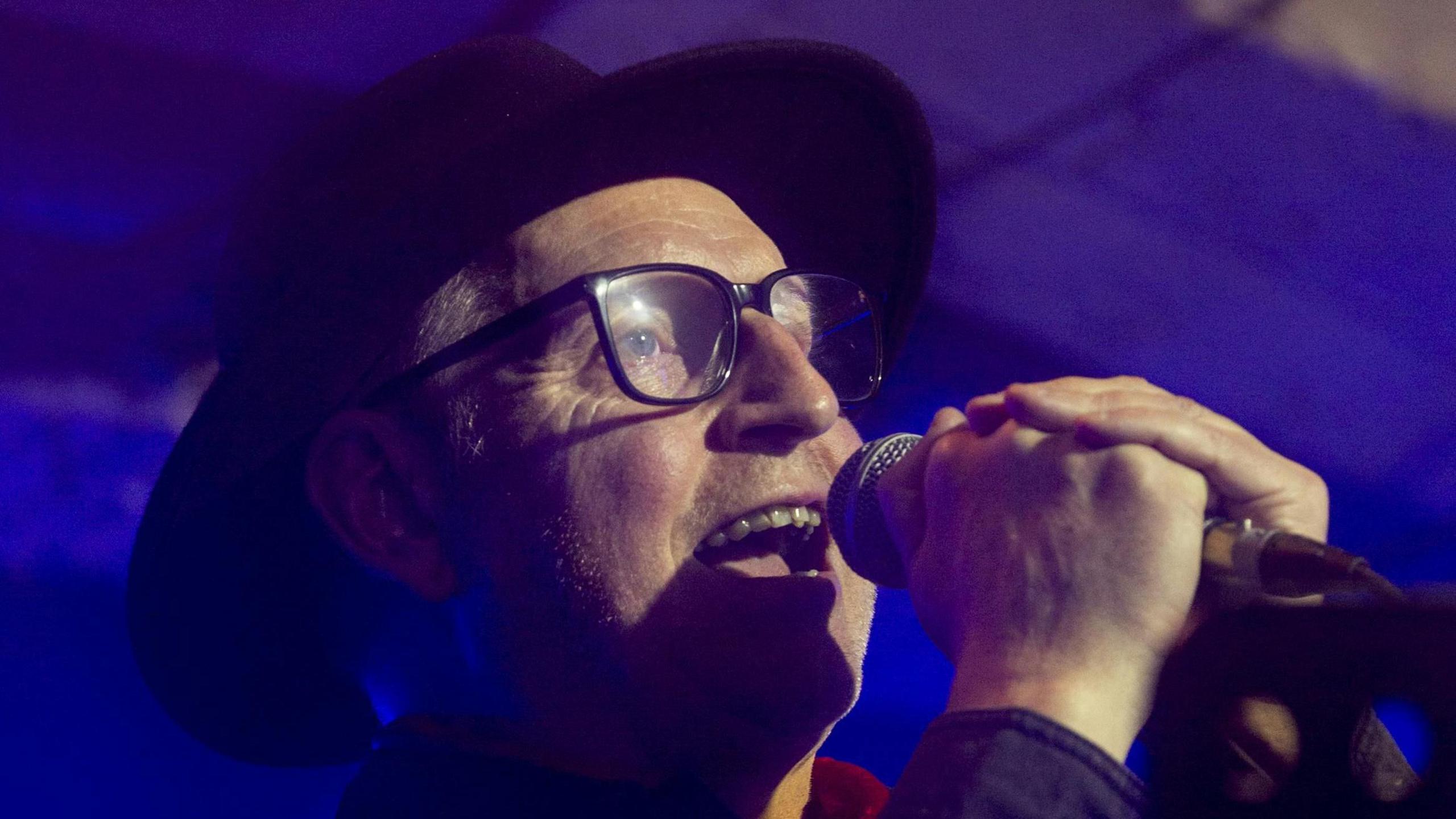 David onstage. He is holding a microphone close to his mouth and singing, and wearing a black hat